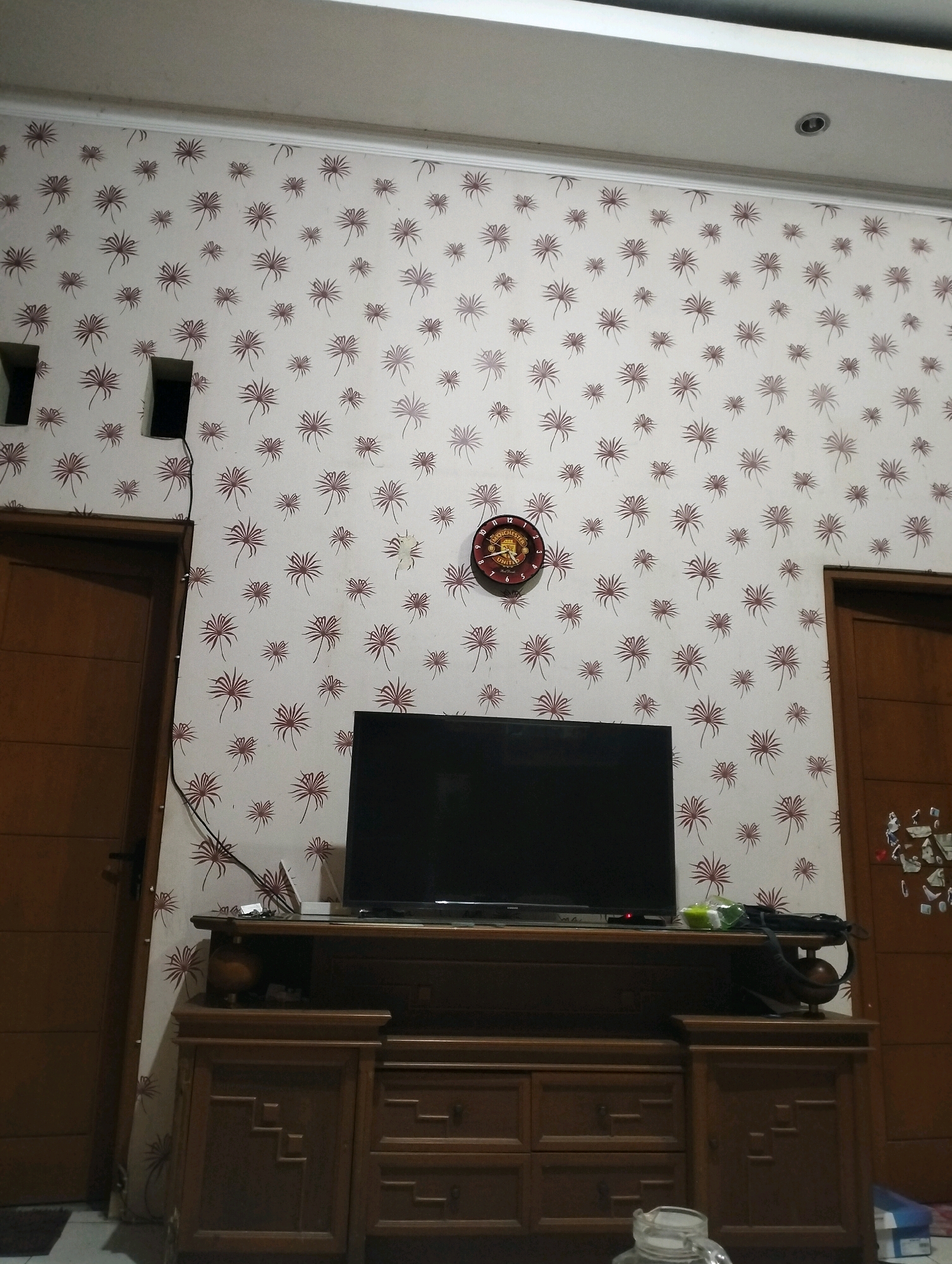 The image depicts an indoor scene, likely a living room, in Sragen, Indonesia. The walls are covered with light-colored wallpaper featuring a repeating pattern of stylized maroon floral or palm motifs. Two wooden doors are visible, one on the left and one on the right, both appearing to be a medium brown color.
In the center of the frame, a black flat-screen television rests on a dark brown wooden entertainment center. The entertainment center has multiple drawers and cabinets with decorative carved detailing. Above the television, on the wall, a round analog clock with a dark red face and white numerals is mounted. A black electrical cord runs vertically down the wall from an unseen source, disappearing behind the entertainment center.
On the left side of the image, near the top, are two dark, rectangular openings in the wall, possibly electrical outlets or vents. The ceiling is visible at the top of the image, with a white molding along the edge of the wall and a circular ceiling fixture.
The overall impression is that of a domestic living space, possibly a bedroom or family room, furnished with functional and decorative elements. The lighting appears to be artificial, creating a warm, somewhat dim ambiance. No people are visible in the frame.