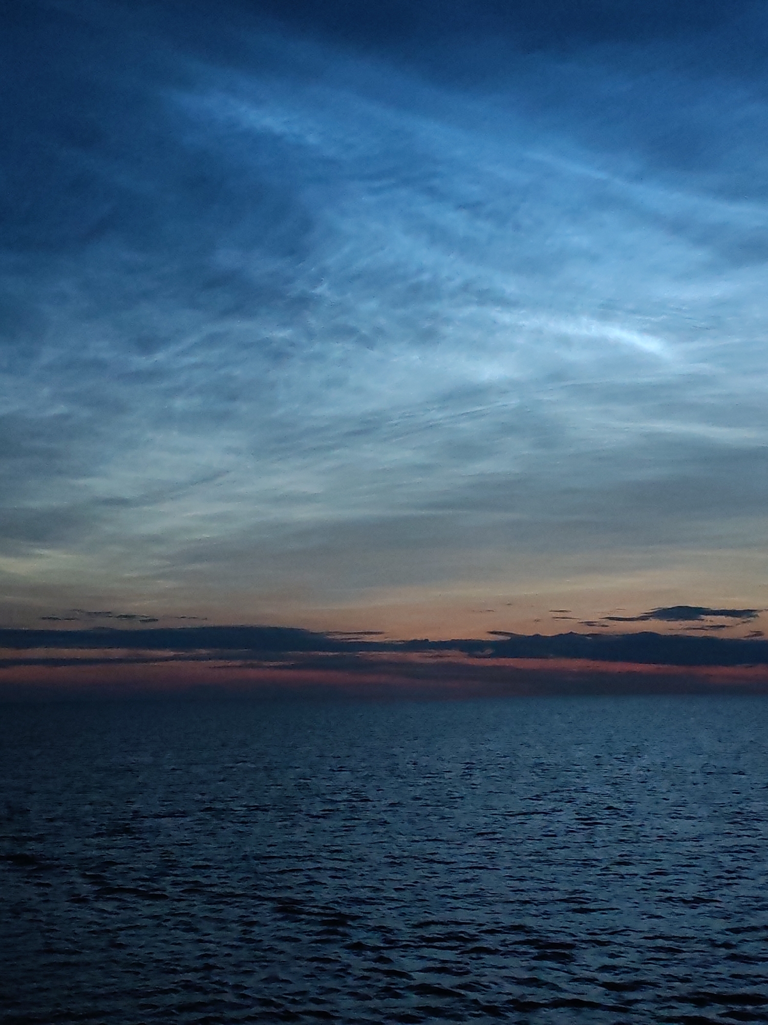 The image depicts a twilight scene over a body of water, likely the sea or a large lake, with a horizon line bisecting the frame. The upper portion of the image is dominated by the sky, which transitions from a deep, dark blue at the top to lighter, more muted tones closer to the horizon. Wispy, luminous clouds, possibly noctilucent clouds, are visible, catching the last light of the sun and appearing as streaks of ethereal white and pale blue against the darker sky.
Below the sky, a thin band of color indicates the sunset. It's characterized by hues of orange and pink, overlaid with darker cloud formations that appear as silhouetted shapes against the fading light.
The lower half of the image is filled with water. The surface is calm but textured, with ripples and small waves visible, reflecting the dim light of the sky and creating a dark, somewhat textured expanse. The overall impression is one of tranquility and the transition from day to night. The image does not contain any people, objects, or discernible human activity, focusing solely on the natural landscape at dusk. The location is specified as Ozernoe Ust'e, Russia, suggesting a coastal or lakeside setting within that region.