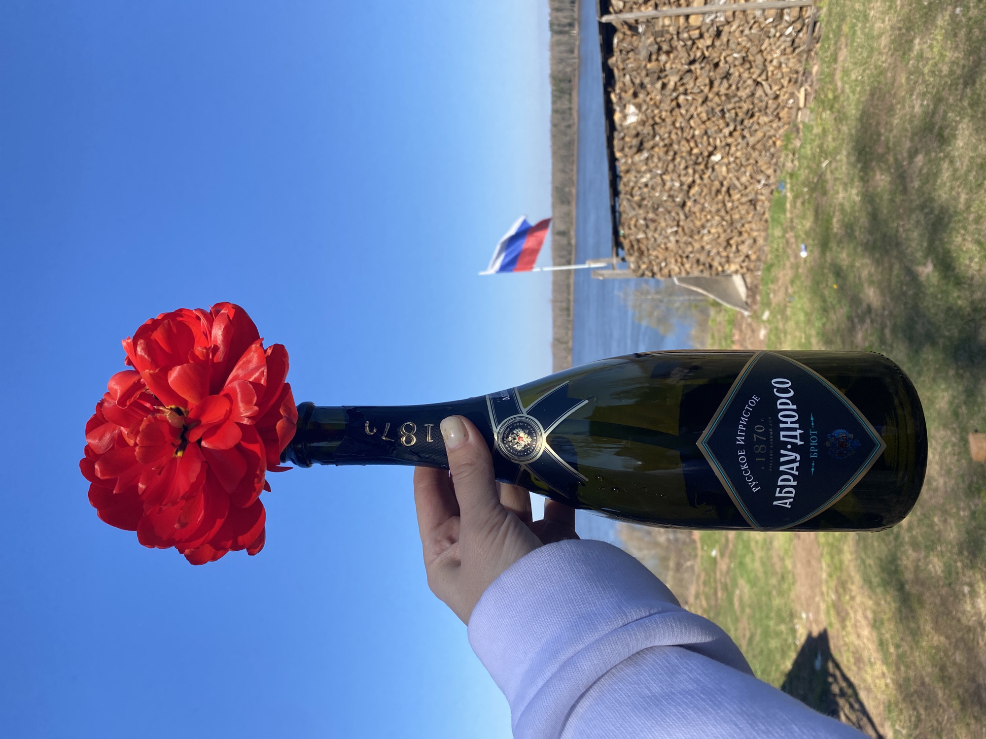 A hand holds a bottle of "Abrau-Durso" sparkling wine (Russian origin) with a red flower inserted into the neck. The background includes a body of water, a Russian flag, and stacked firewood, suggesting a riverside or lakeside location, with grass and blue sky also visible. The image location is Ольховка, Russia.