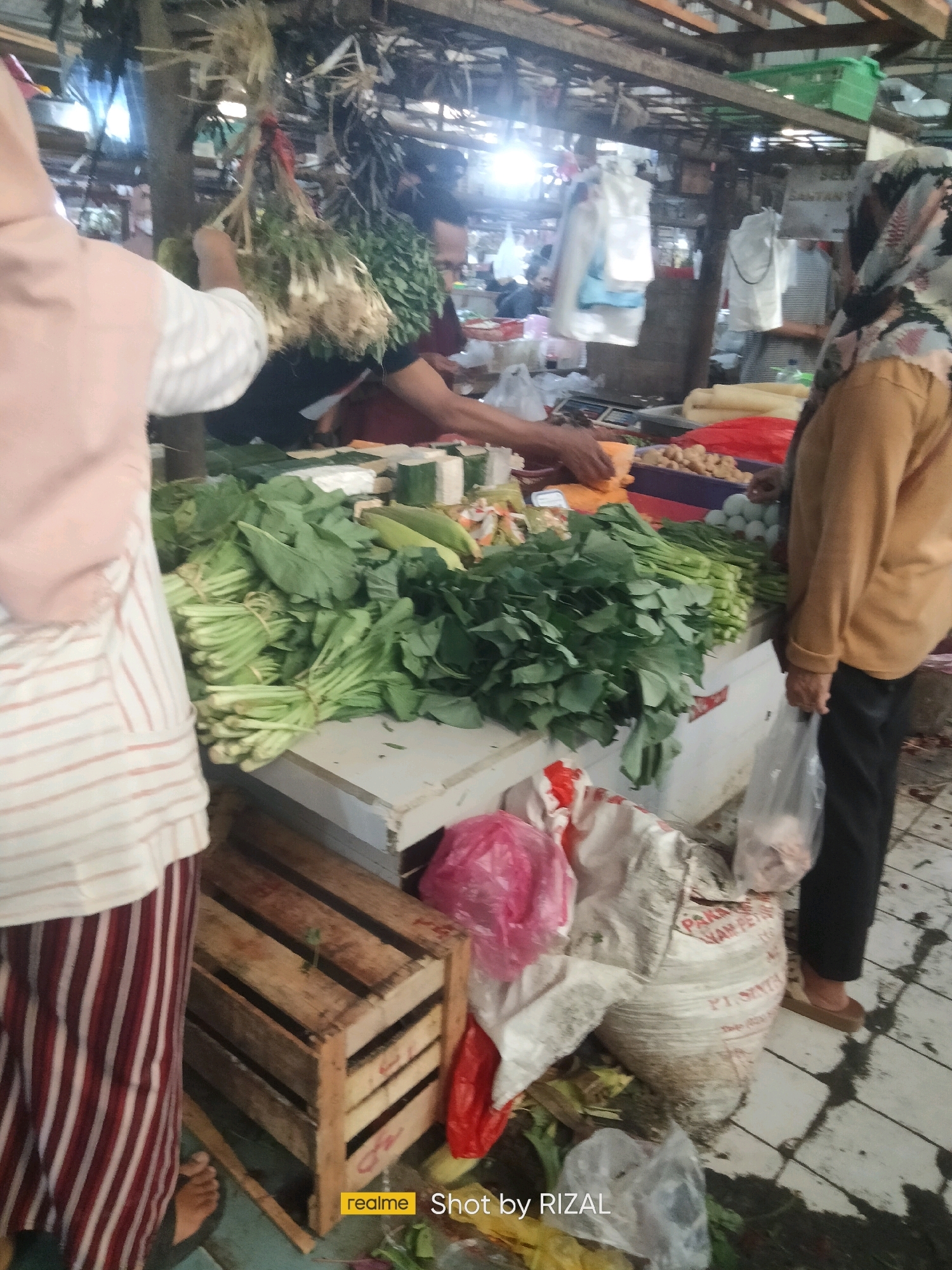 The image depicts an outdoor market scene, likely in Bojong Nangka, Indonesia. In the foreground, a person wearing a pink hijab and a striped skirt stands to the left, facing away from the camera. In the center, a stall is laden with fresh produce, including bunches of leafy green vegetables and what appear to be bundles of scallions or similar herbs hanging from above. To the right, another person wearing a yellow top and headscarf is visible, holding a plastic bag. Various bags and crates are scattered around the stall, and the ground is paved with tiles. The overall environment suggests a busy marketplace with a focus on fresh food.