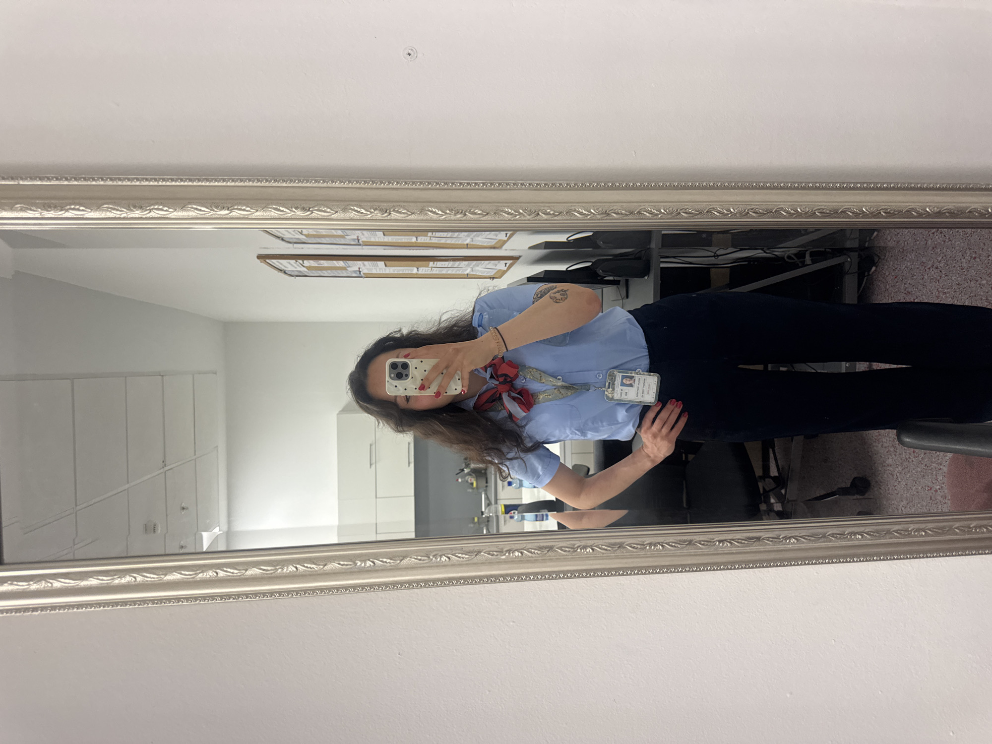 A woman is taking a mirror selfie indoors. The mirror has a silver frame. The woman is wearing a light blue button-down shirt, a red and patterned scarf, and dark pants. She has a name tag on her shirt. A tattoo is visible on her left arm. The background shows office equipment and a desk area. A sign is visible on the wall in the reflection. The location appears to be in a workplace; however, there are no details in the picture that would indicate it is Burgas, Bulgaria.