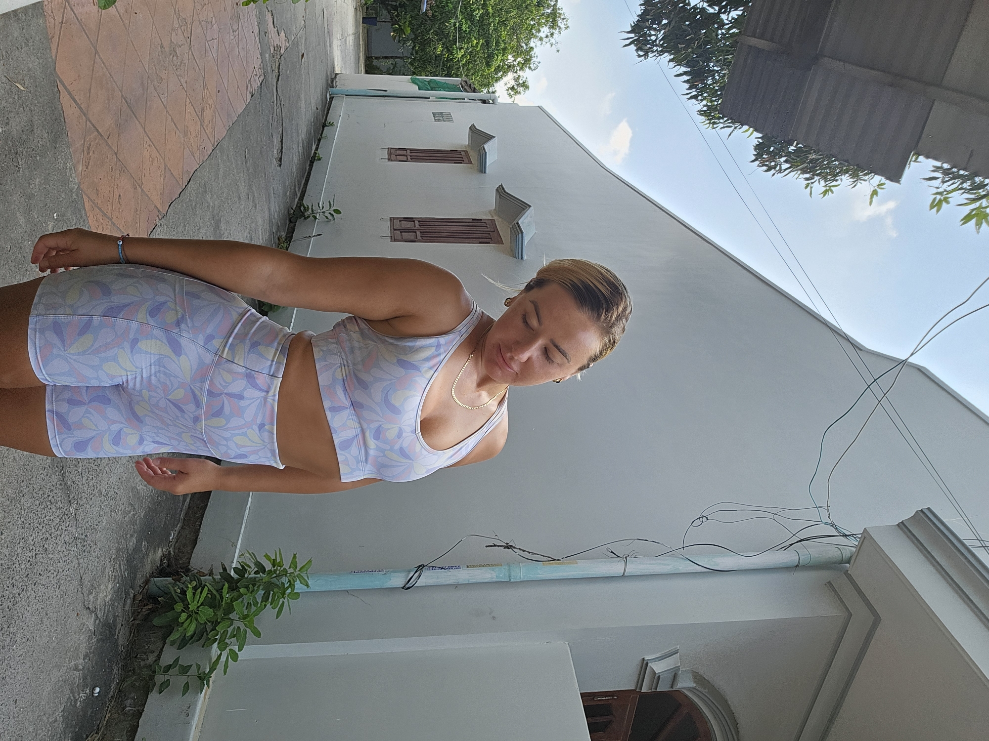 A young Caucasian woman with blonde hair pulled back is standing outdoors in front of a light-colored building with a curved wall. She is wearing a matching athletic set: a sports bra and high-waisted shorts, both featuring a pastel floral print. Her head is tilted down and to the side, with her eyes closed. The environment appears to be an alleyway or side street, with a tiled walkway on the left and a concrete surface on the right. Trees and foliage are visible in the background. The architecture suggests a location in Phu Thuy Ward, Vietnam, with details like the building's windows and external piping being characteristic of the region. The lighting indicates it is daytime, with a bright sky.