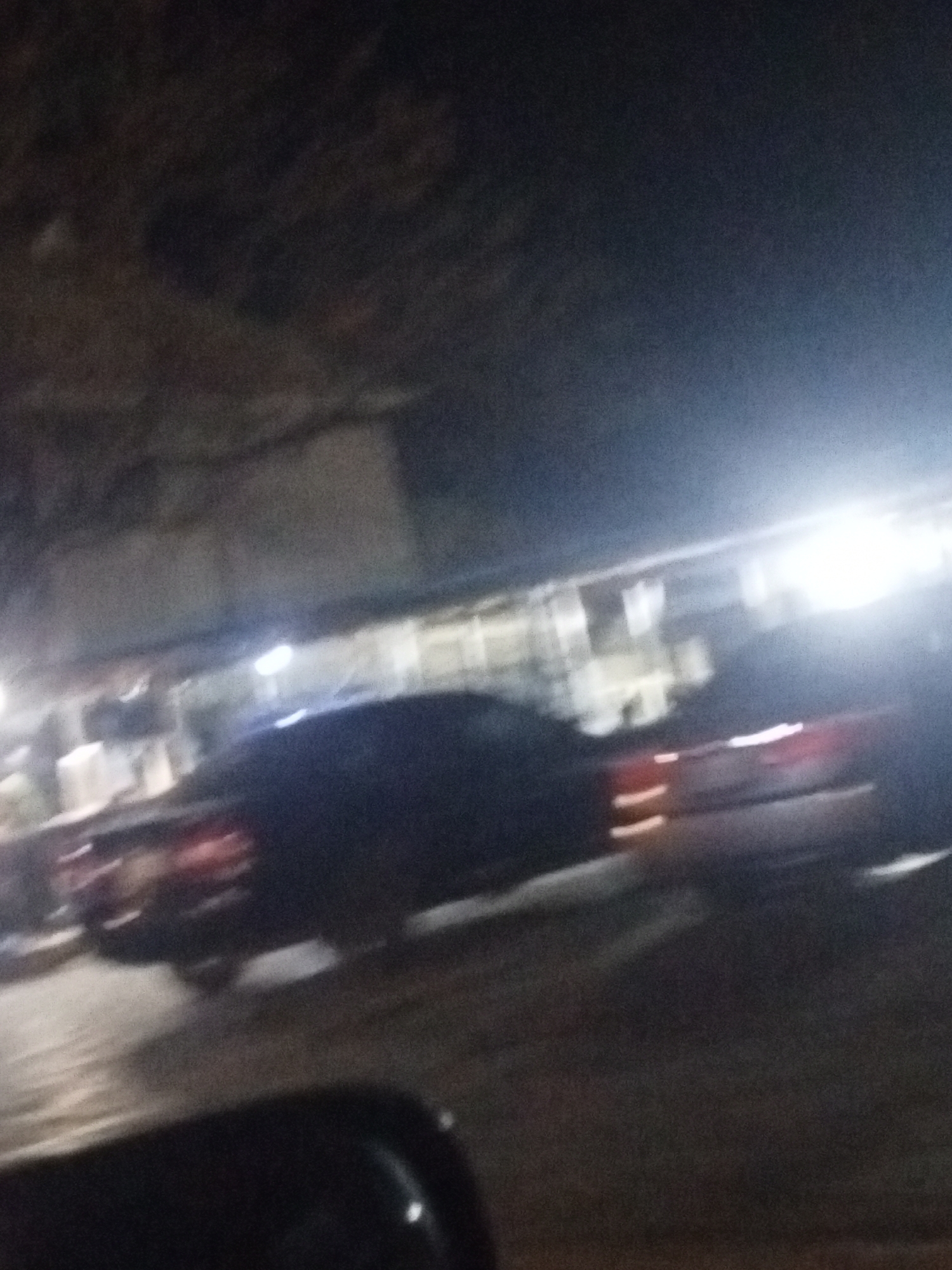 The image shows a dark street scene at night in an unknown city. There are cars visible, including at least one dark-colored sedan with its taillights illuminated. Streetlights are present, casting bright glows on the street and nearby buildings. The foreground is out of focus, suggesting the photo was taken from inside a vehicle. Buildings are visible in the background, but their details are obscured by the darkness and light. The overall impression is one of urban night life, with movement and artificial illumination.
