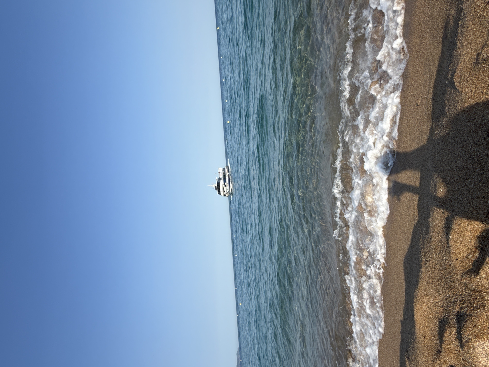 This is a vertical, wide-angle photograph of a beach scene. The image is oriented sideways. On the left side of the image, a vast expanse of clear blue sky dominates. To the right of the sky is the calm, blue water of the sea, which gradually becomes more transparent as it approaches the shore. A small white boat with a black top is visible in the distance, floating on the water. A line of yellow buoys stretches across the sea, marking a boundary.
The foreground of the image features a sandy beach. The sand is fine-grained and has a light brown color. The edge of the water is visible, with small waves lapping at the shore, creating white foam. The right side of the beach shows the shadow of a person, casting a long silhouette across the sand. The shadow suggests that the sun is low in the sky, indicating either early morning or late afternoon. The overall mood of the image is peaceful and serene, evoking a sense of a relaxed beach day. The location is identified as Blanes, Spain. There is no visible text in the image.