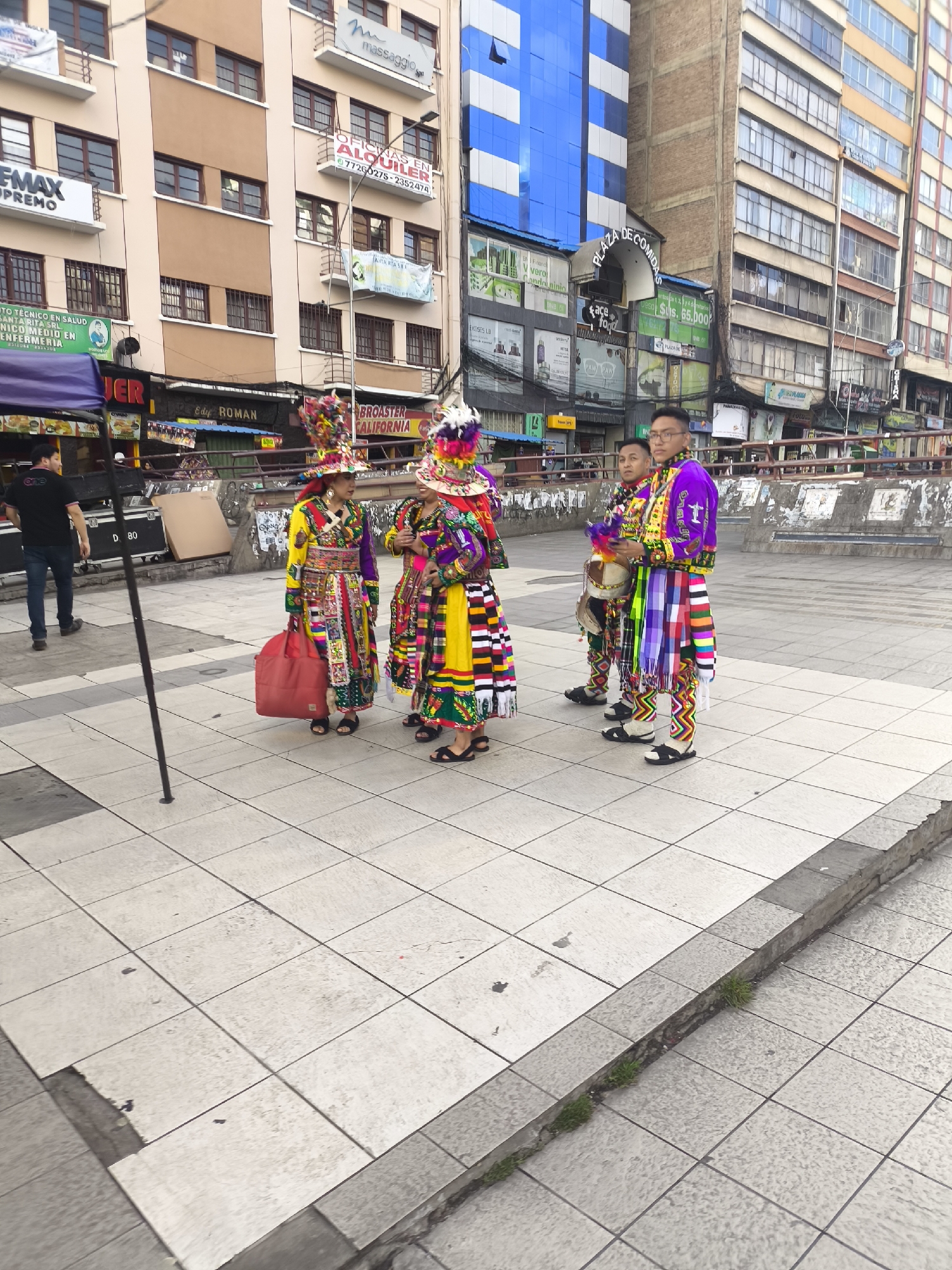 The image depicts an urban street scene in La Paz, Bolivia, situated in front of multi-story commercial buildings, including one labeled "PLAZA DE COMIDAS." Four individuals are prominently featured in colorful, elaborate traditional Bolivian costumes. On the left, two women face each other, possibly conversing; both wear long, multi-patterned dresses and ornate feathered or decorative headwear. One woman carries a red bag. To their right, two men stand, similarly dressed in vibrant, patterned garments, including striped elements and patterned pants; one man holds a small drum. A fifth individual, a man in a black t-shirt and jeans, walks away from the group on the far left. The ground is paved with light-colored square tiles, showing signs of wear. A low concrete barrier with some graffiti and sporadic grass growth separates the immediate paved area from a lower ground level. The background features various commercial buildings with multiple windows, balconies, and visible signage in Spanish, including "OFICINAS EN ALQUILER" (Offices for Rent) and "Edif ROMAN." A building with a modern blue reflective facade is visible in the middle-right background.