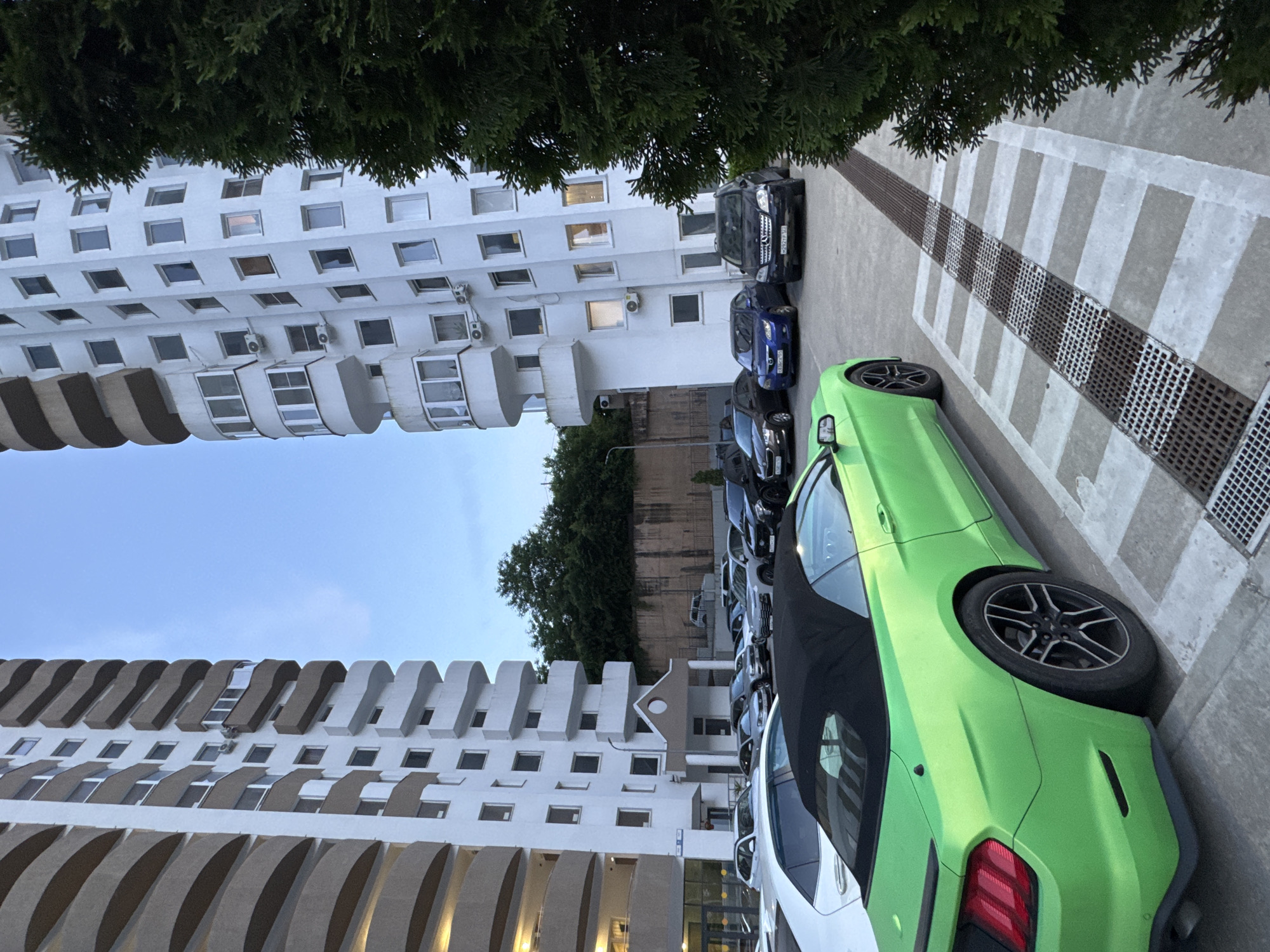 The image depicts a parking lot in front of a modern apartment building in Sochi, Russia. The building features a curved facade with multiple balconies. Several cars are parked in the lot, including a bright green convertible in the foreground, a black Mercedes-Benz, and a blue sedan. A large evergreen tree is visible on the right side of the frame, partially obscuring the building. The sky is overcast, suggesting it is either early morning or late evening. The overall scene is urban and residential.