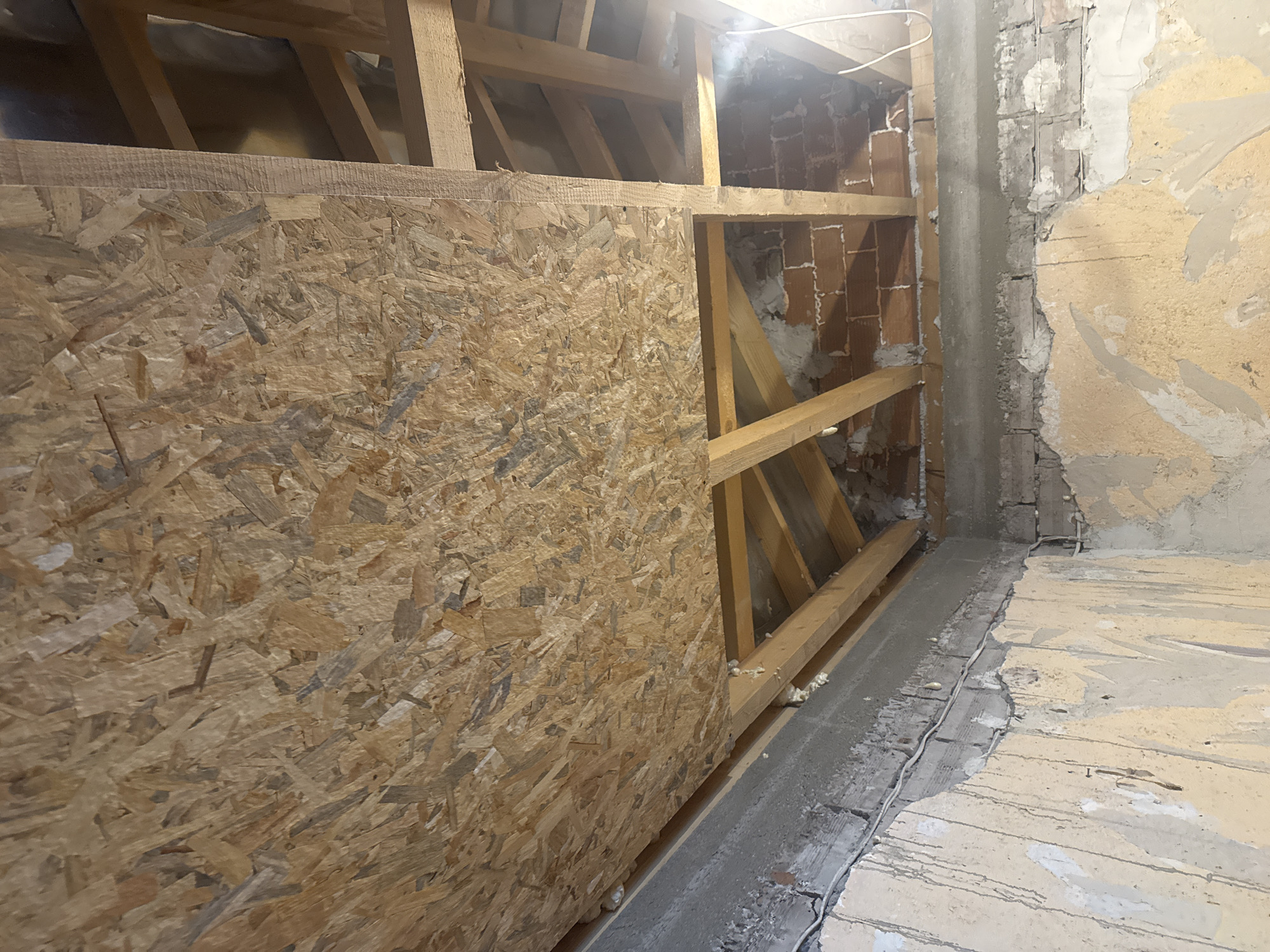 The image depicts an interior space in Sarnevo, Bulgaria, undergoing construction or renovation, captured from a low-angle perspective looking upwards. The ceiling is partially covered with light brown Oriented Strand Board (OSB) panels, characterized by visible wood chips and strands. Exposed wooden rafters and beams form a structural framework beneath and adjacent to the OSB panels. In the upper right and lower right sections, portions of the ceiling or upper wall reveal exposed red brickwork and additional wooden beams. White expanding foam insulation is visible at the junction of the OSB and the adjacent wall, as well as on some of the wooden supports. A single white electrical wire descends from the upper right corner, with a bright light source creating a glare in that area.
The walls in the lower half of the image show multiple layers of material. The lowest visible layer consists of discolored, peeling yellowish-beige paint or wallpaper, revealing a lighter substrate beneath. Above this, a rough, grey concrete or plaster band runs horizontally, beneath which additional exposed brick is visible. White electrical wiring is routed along these wall sections, both on the surface and partially embedded. No individuals or specific actions are present within the frame. The overall environment indicates an unfinished or deconstructed interior space.