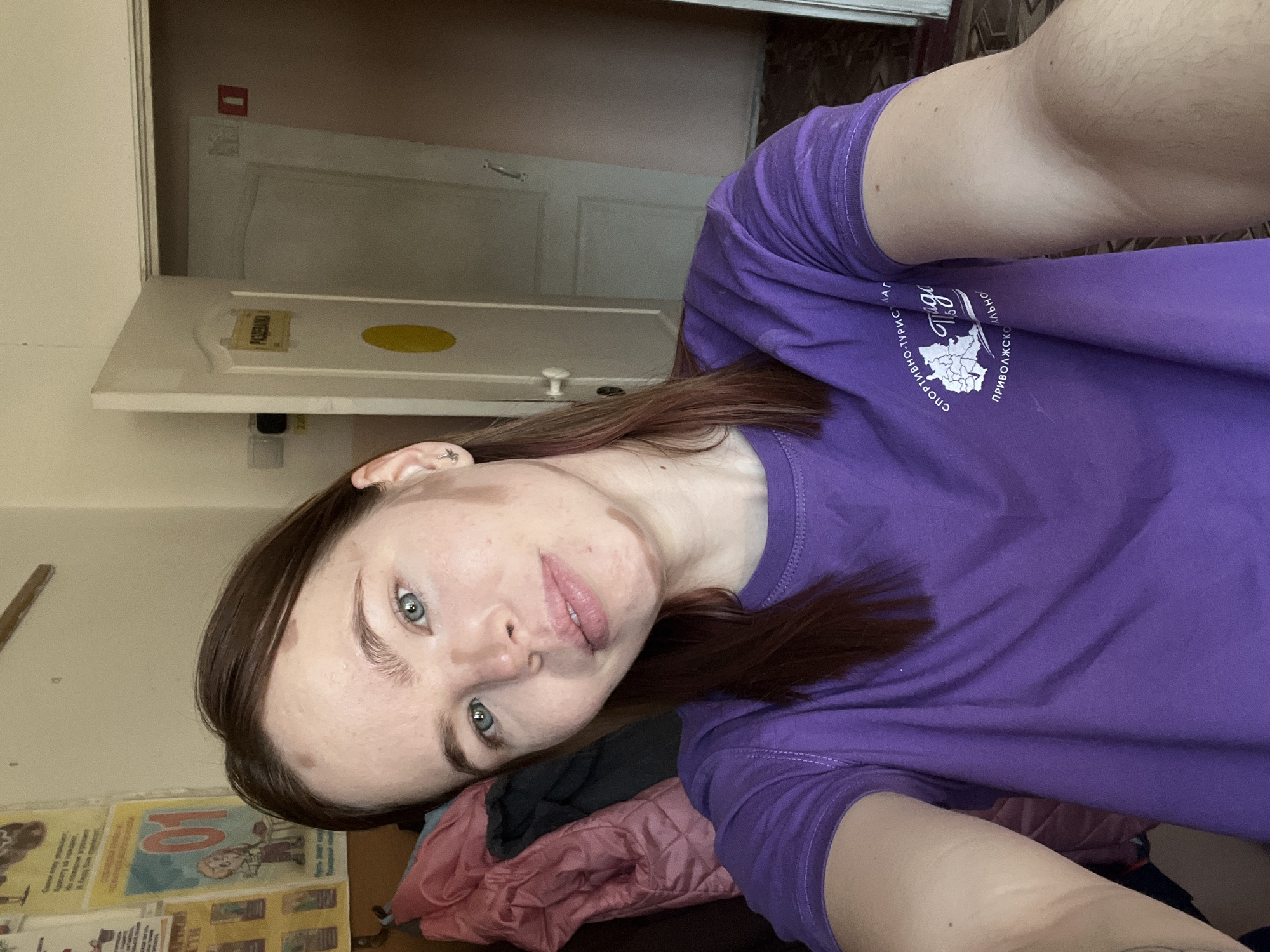 A selfie of a fair-skinned person with auburn hair and blue eyes, wearing a purple t-shirt with Cyrillic text and a landscape illustration, is oriented diagonally. The background appears to be an interior space with a partially opened white door, a bulletin board with posters and papers, and folded clothing. The location is Unknown, Unknown.