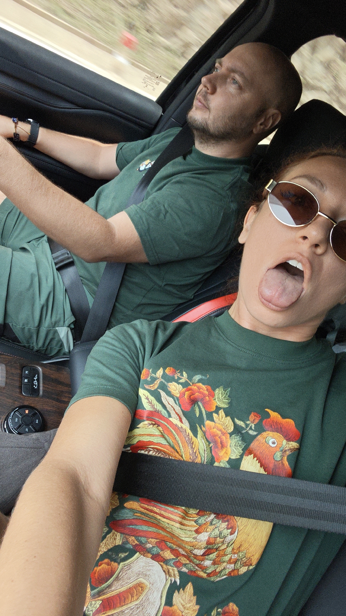 A selfie taken from inside a moving car captures two individuals, a woman in the foreground and a man driving, presumably in Duong Quy, Vietnam.
The woman, positioned in the front passenger seat, holds the camera, sticking her tongue out playfully. She wears dark, round sunglasses and a dark green t-shirt adorned with a large, colorful embroidered pattern featuring a rooster and various floral elements. A black seatbelt is fastened across her chest. Her arm is visible in the lower left of the frame.
The man, driving the vehicle, is seen in profile, looking straight ahead, appearing focused or contemplative. He has a shaved head and a short beard, wearing a plain dark green t-shirt and matching dark green pants. A black seatbelt crosses his chest. His left arm, adorned with a black watch or bracelet, rests near the steering wheel.
The car's interior features dark upholstery and dashboard elements, with a distinct wooden trim visible on the center console, which houses various control buttons and a rotary knob. The background outside the car is blurred, indicative of movement, showing hints of green foliage and a road or natural scenery. The scene appears to be taking place during daylight hours. Both individuals are dressed in coordinating dark green attire.