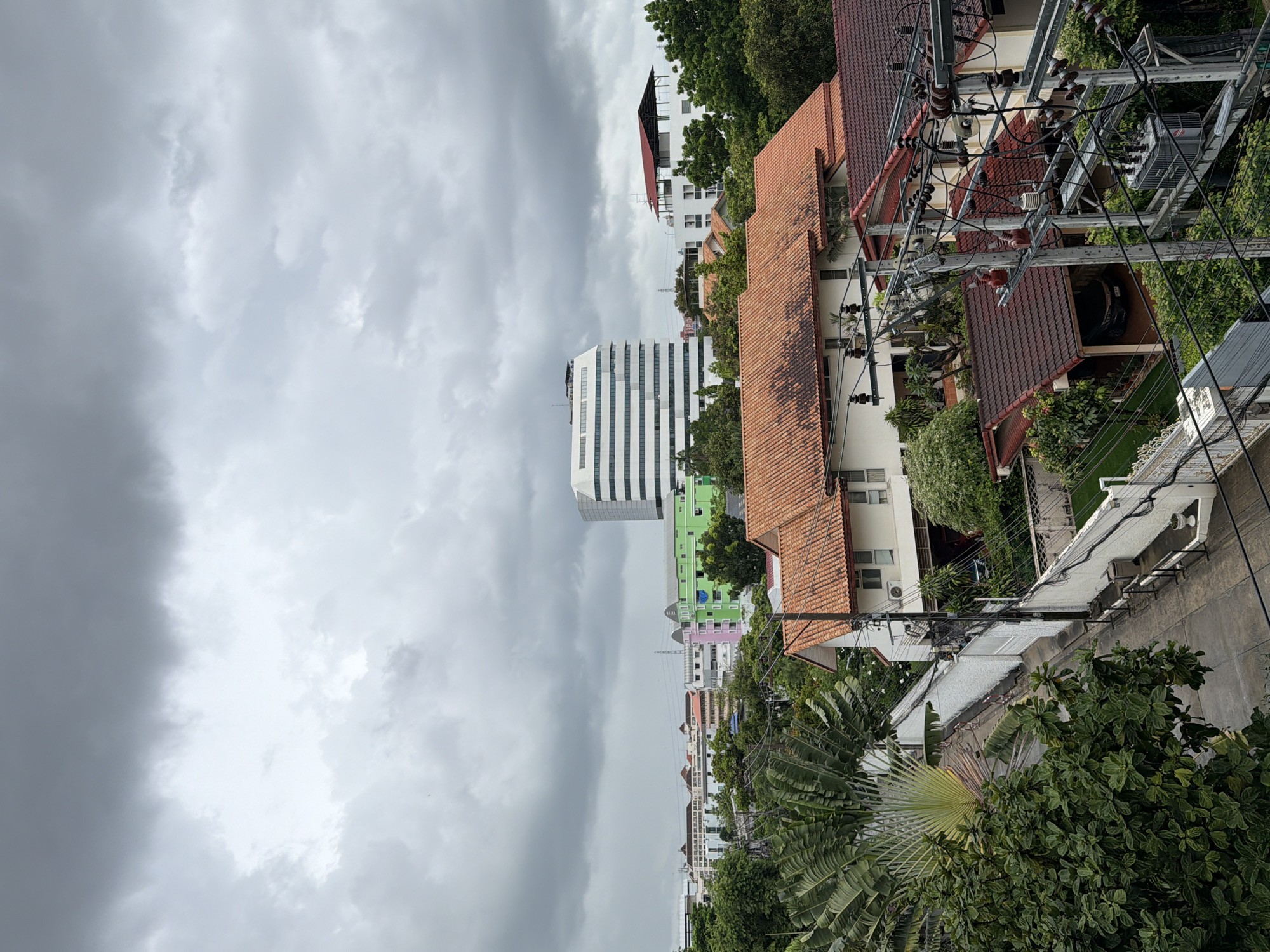The image shows an overcast sky over a city with a mix of residential and commercial buildings. In the foreground, there are several houses with red tile roofs and lush green trees. A street runs between the houses, lined with white walls and electrical poles with transformers and numerous wires. In the background, there are taller buildings, including a white, curved multi-story building with many windows, and other structures in various colors like green and pink. The overall scene suggests a dense urban environment under a cloudy sky, likely in Bangkok, Thailand, given the architectural style and vegetation. There are no people visible in the image.
