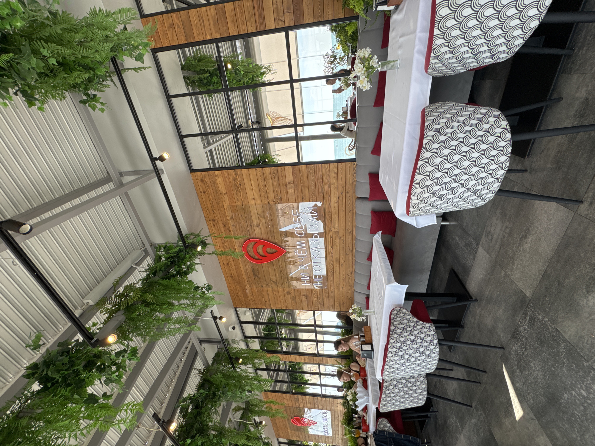 The image depicts the interior of a restaurant in Sochi, Russia, with a view of the Black Sea through large windows. The restaurant features a modern design with a wooden accent wall adorned with a red heart-shaped logo and Russian text that translates to "Don't deny yourself anything." Hanging plants cascade from the ceiling, adding a touch of nature to the space.
Several tables are set with white tablecloths, and guests are seated at some of them, including two women in the foreground. The flooring is a dark, textured tile. The overall ambiance suggests a bright and airy dining experience, possibly during the daytime, given the natural light entering the restaurant.