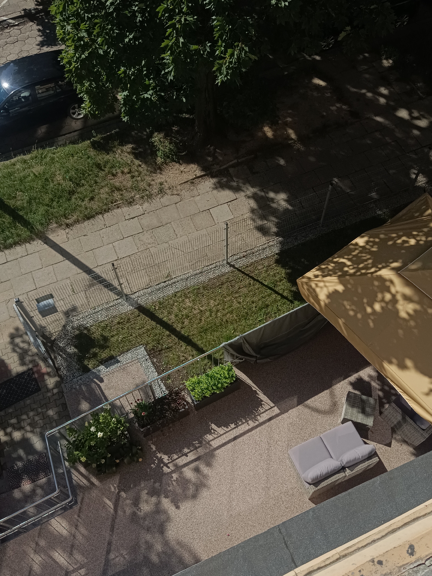 The image shows an aerial view of a residential building's exterior area in the city of Łódź, Poland. In the upper left, a black car is parked on a paved area with some grass next to it, bordered by a sidewalk. A large tree with dense green foliage casts dappled shadows across the scene. Further down, a metal fence separates a grassy area from a paved walkway. Adjacent to the walkway, a patio area is visible with outdoor furniture, including a light brown umbrella, a two-seater grey sofa with cushions, and two grey chairs. Planters with green foliage are positioned along the edge of the patio. The building's facade and a balcony railing are also present in the foreground. The overall scene depicts a sunny day with strong shadows, suggesting a well-maintained outdoor space.