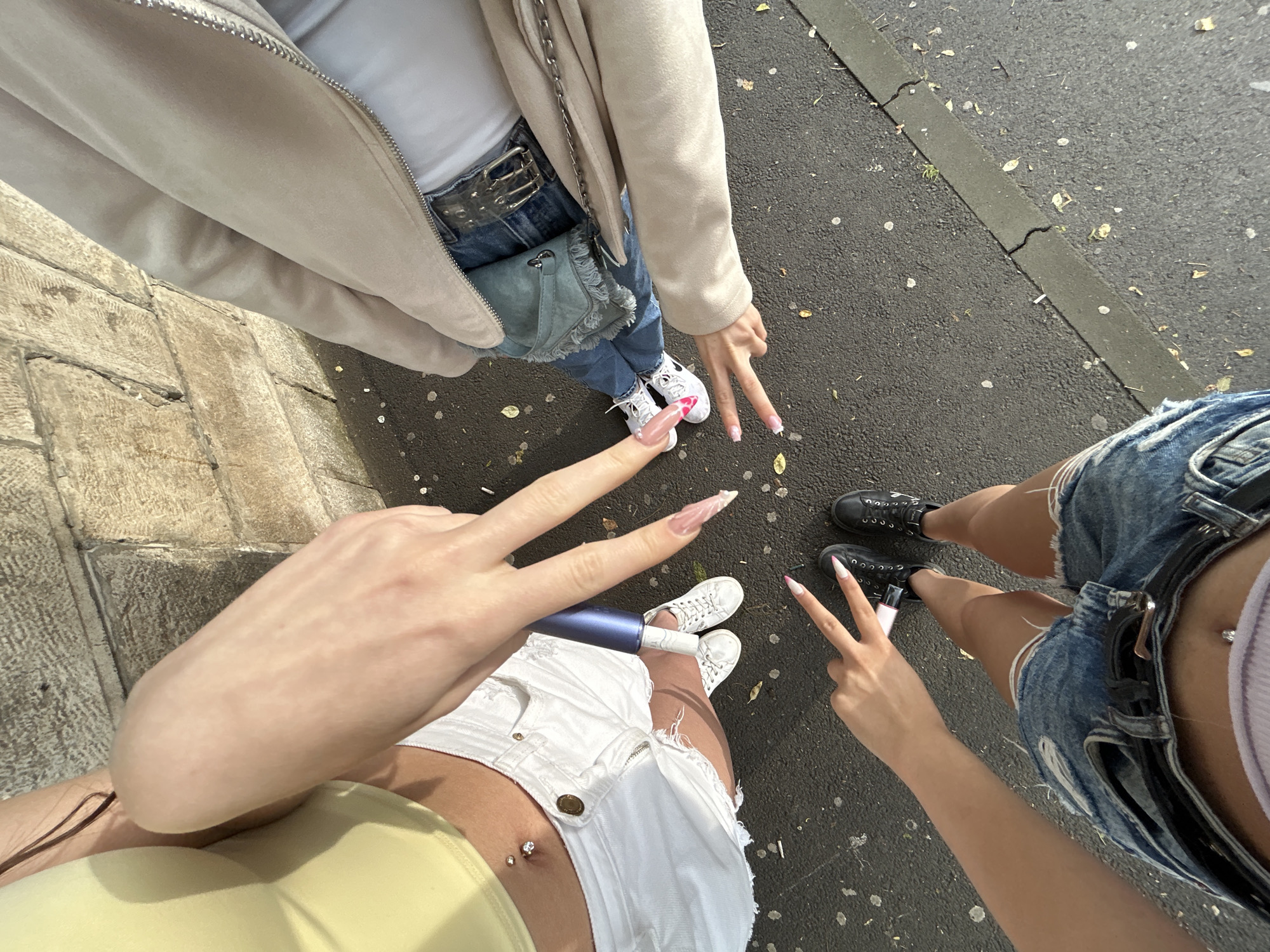 Three individuals are captured in a high-angle, close-up shot, standing on a paved surface, possibly a sidewalk or street, with a stone wall in the background. The environment appears to be outdoors, with scattered leaves and small debris on the ground.
The person on the left is wearing a light yellow cropped top and white ripped denim shorts, with a belly button piercing visible. They are wearing white sneakers. One of their hands is prominently displayed in the foreground, making a peace sign with long, manicured fingernails. A blue cylindrical object, possibly a vape pen or cosmetic item, is held near their shorts.
The person in the middle is partially visible, wearing a light beige jacket zipped over a white top and blue jeans. They are wearing white sneakers, similar to the person on the left.
The person on the right is wearing dark denim shorts with a black belt and a white top. They are wearing black lace-up shoes. One of their hands is also visible, making a peace sign with manicured fingernails.
The lighting suggests it is daytime, with sunlight casting shadows and highlighting the textures of the stone wall and the clothing. The image focuses on the lower bodies and hands of the individuals, emphasizing their fashion choices and the gestures they are making. No specific landmarks identifiable as being in Shumen, Bulgaria, are present in the image.