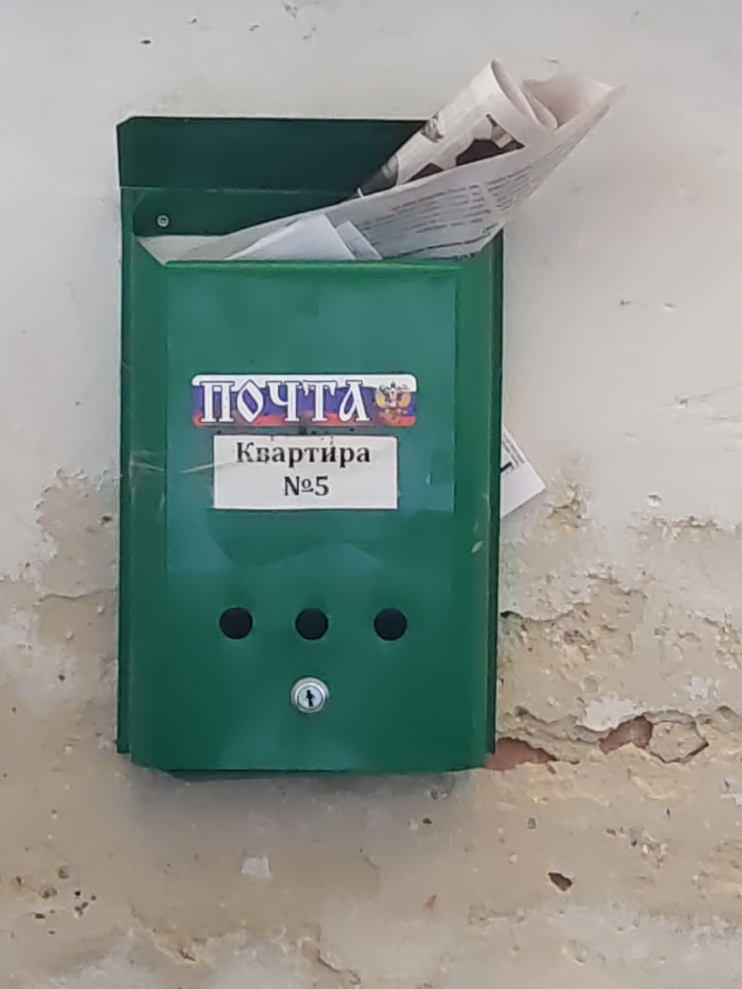 A green metal mailbox is attached to a light-colored, weathered wall. The mailbox has a label with Russian text, "ПОЧТА" (POCHTA, meaning "POST") and "Квартира №5" (Apartment No. 5). A newspaper or a bundle of papers is sticking out of the top of the mailbox. Three black circular holes are visible on the front of the mailbox, with a small keyhole below them. The wall shows signs of wear and tear, with peeling paint and textured patches. The scene is located in Pukhlyakovskiy, Russia.