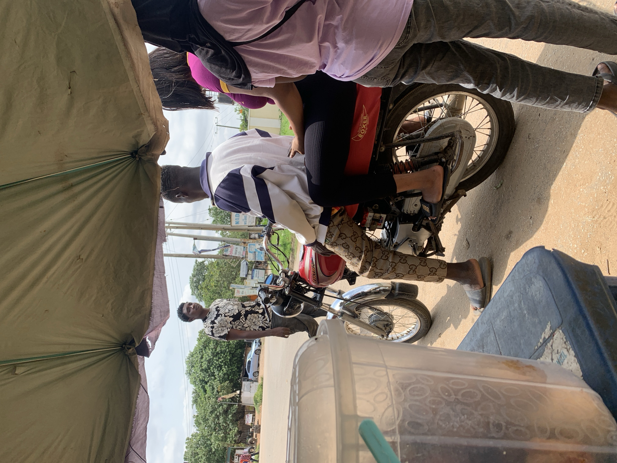 Two people are on a motorcycle, with one person in the front driving and another person behind them. The person in front is wearing a white and blue jacket and a patterned shirt underneath. They are looking down at their hands. The person in the back is wearing a pink and purple shirt and black pants. They are leaning forward and appear to be holding onto the driver. The motorcycle has a red fuel tank with the word "Boxer" written on it in white text. In the foreground, there is a large, olive-colored tarp or umbrella, and a blue plastic container. A third person is standing to the left of the motorcycle, wearing a black and white patterned shirt and dark pants. They are looking towards the camera. The scene appears to be outdoors, with trees and a road visible in the background. There are also some signs and poles in the distance. The lighting suggests it is daytime, possibly with some shade provided by the tarp. The overall atmosphere seems casual, and the people appear relaxed. It's possible they are traveling or taking a break. Some street signs are visible in the background, and given the location context of Abuja, Nigeria, this is likely a street scene within the city.