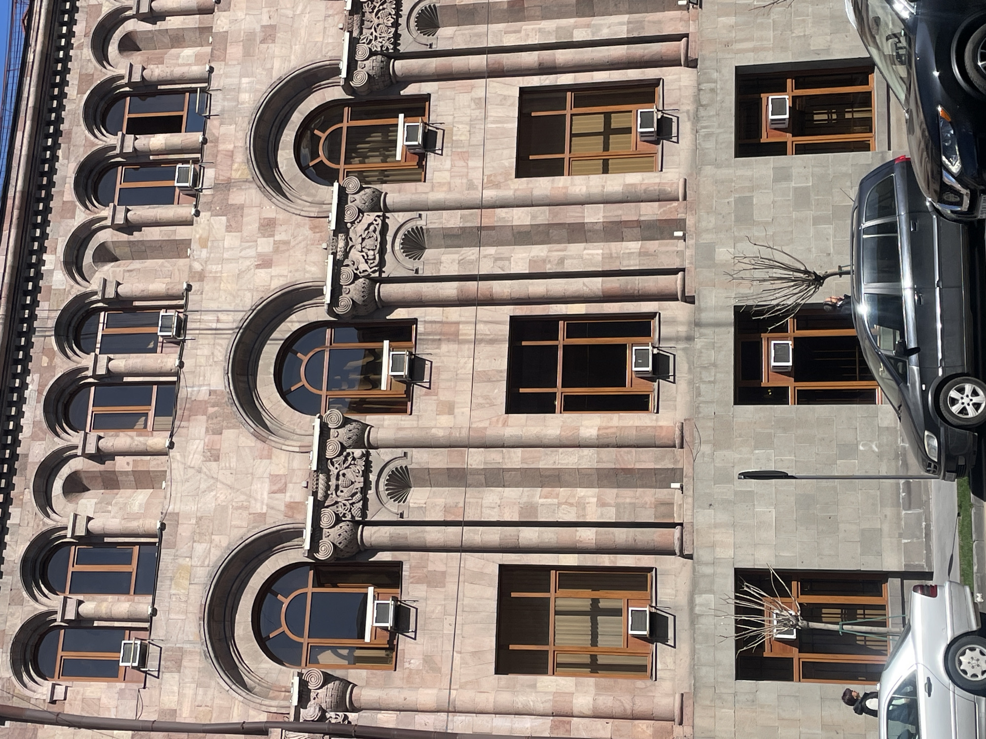 The image shows a building facade in Yerevan, Armenia, constructed with a light brown stone. The building features a pattern of arched and rectangular windows framed in wood. Air conditioning units are visible on the outside of many of the windows. Columns separate the window groupings, adorned with ornamental details. The building's right side contrasts with gray brick construction. Several cars are parked along the street, partially obstructing the view of the building's lower floors. A bare tree stands in front of one of the windows on the right. A single street light stands in the background. A person is seen near the white car.