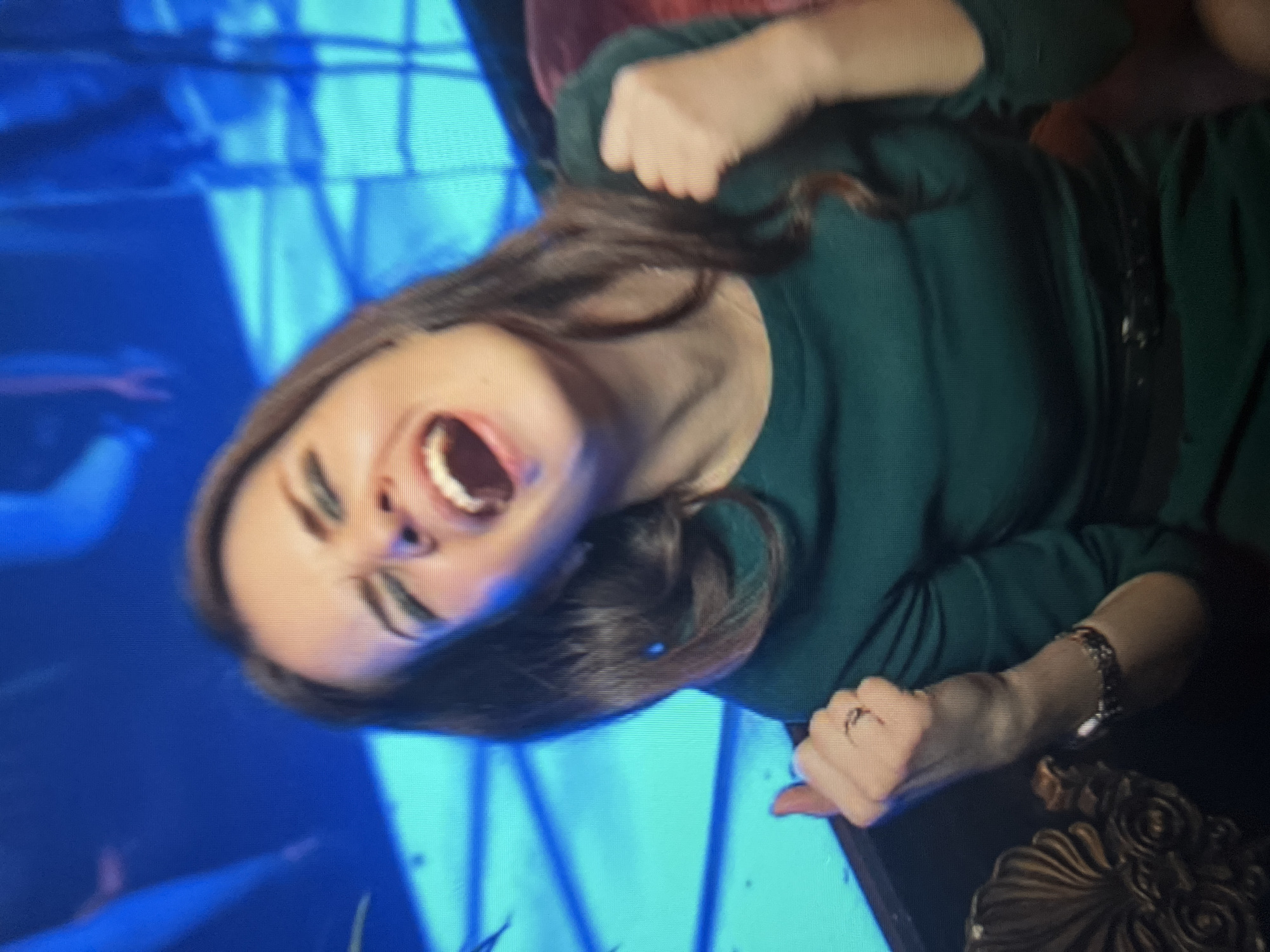 A woman with long brown hair and green eye makeup is shown from the chest up. She is wearing a dark green dress and has her mouth open in a shout or cheer. Her right arm is raised with her fist clenched, and her left arm is also raised with her thumb up. A ring is visible on her left hand, and a bracelet is on her right wrist. The background is blurry and appears to be lit with blue lights, suggesting an indoor event such as a concert or party. The image's pixelated quality indicates it was likely taken with a low-resolution camera or from a distance. There is no indication of the location being the city of Qarağandı city administration, Kazakhstan.
