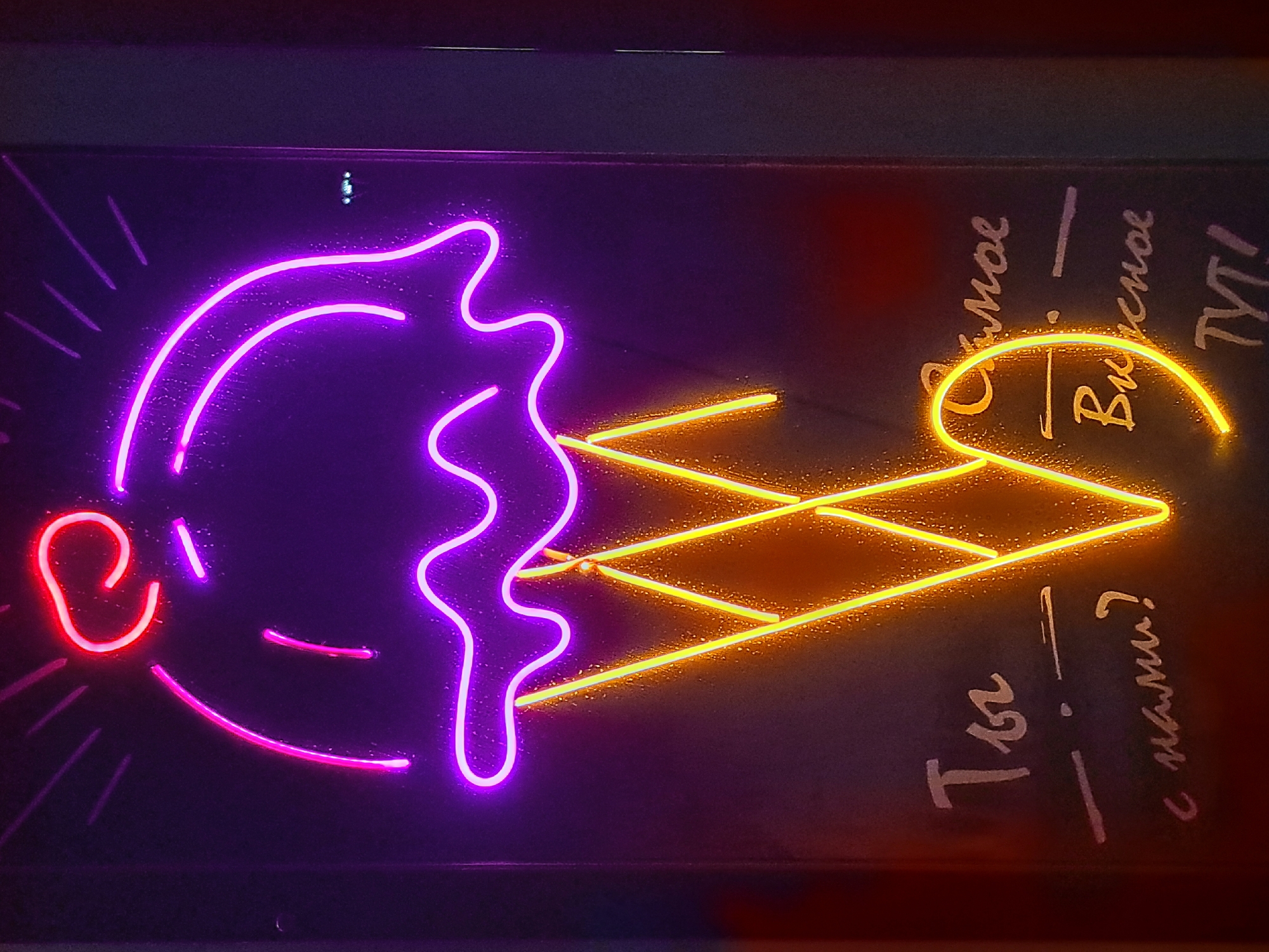 A neon sign depicting an ice cream cone is displayed vertically. The ice cream portion is purple with pink and red accents at the top, resembling a cherry. The cone is outlined in bright yellow neon. Below the cone, white lettering is visible against the dark background. The text reads, "Ты - с нами? Самое вкусное ТУТ!". This translates to "You - with us? The most delicious is HERE!". The sign appears to be illuminated in a dimly lit environment, possibly a shop or establishment. The location of this sign is confirmed to be Irkutsk, Russia, a city in Siberia.