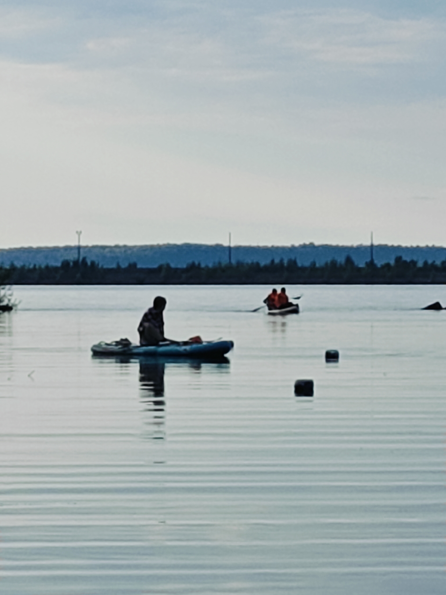 The image depicts a serene, overcast day on a body of water, likely a river or reservoir, given the context of Chaikovsky, Russia, a city situated on the Kama River. In the foreground, a person is seated on a blue kayak, facing away from the camera. Their attire is indistinct due to the distance and lighting, but they appear to be wearing a patterned shirt. The kayak is partially submerged, indicating it is in the water.
Further in the mid-ground, another watercraft, possibly a canoe, is navigating the water. Two individuals are visible in this boat, both wearing bright orange life vests, suggesting safety precautions are being taken. One individual appears to be paddling.
The background is characterized by a hazy, treed shoreline and a pale, cloud-filled sky. The water is calm, with faint ripples reflecting the subdued light. Several dark, rectangular objects are floating on the surface of the water at various distances, their purpose unclear. The overall impression is one of a tranquil outdoor activity in a natural setting.