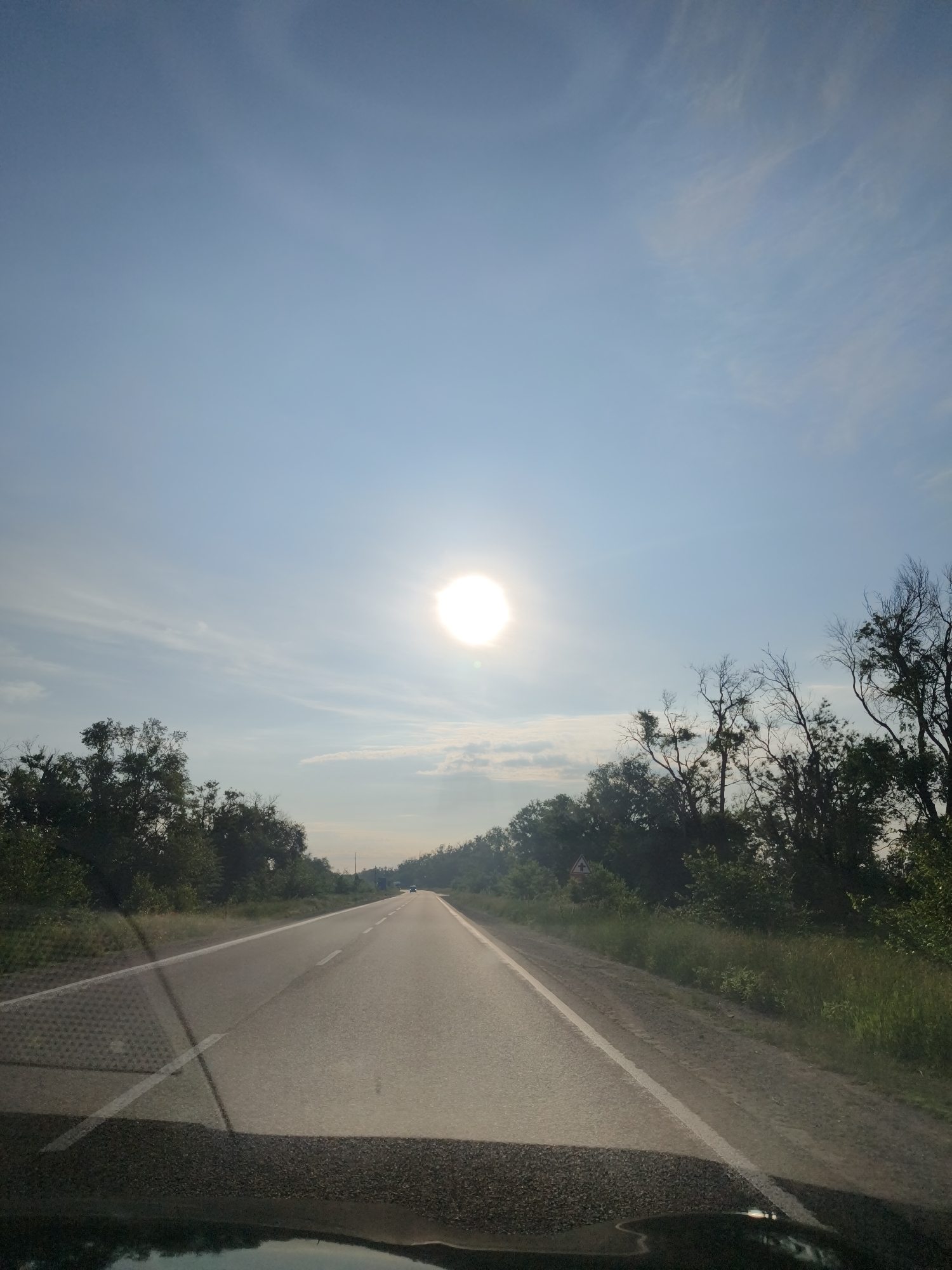 The image presents a forward-facing view from inside a vehicle traveling along a straight, two-lane asphalt road. A brilliant, intense sun is centrally positioned in the upper portion of the clear blue sky, which features scattered, wispy white clouds.
Dense green trees and bushes line both sides of the road, forming a natural corridor. Some trees on the right side appear to have bare branches, contrasting with the lush foliage. The road itself is marked with a solid white line on the right shoulder and a broken white line dividing the two lanes. Further in the distance, the faint outline of another vehicle can be seen on the road ahead.
In the foreground, reflections and shadows from the vehicle's interior are visible on the windshield, including a distinct patterned texture on the left side, possibly from a dashboard mat or object. Partially obscured by the trees on the right side of the road is a yellow, triangular road sign with a red border, likely indicating a warning.
The scene depicts a journey under bright daylight conditions, suggesting it is mid-day or late afternoon given the sun's high position. The weather is clear and fair. The setting is a rural or semi-rural highway in Sofiivka, Ukraine. No explicit text is visible within the image.