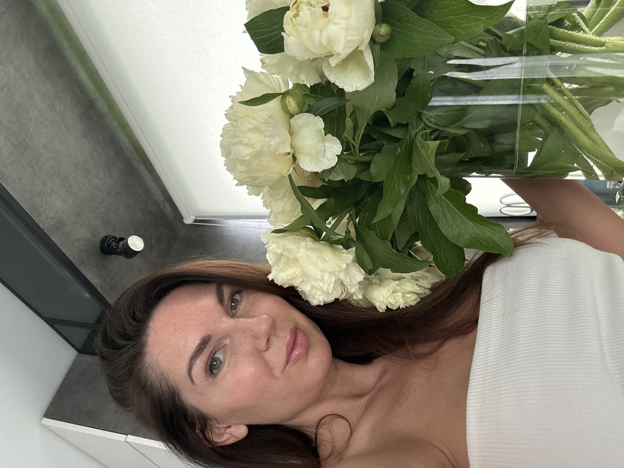A woman is holding a vase full of white peonies with green foliage. She is wrapped in a white towel. The setting appears to be indoors, possibly a bathroom, with light grey and white walls. A light fixture is visible in the top left corner. There is no information to confirm that this image was taken in Chaikovsky, Russia.