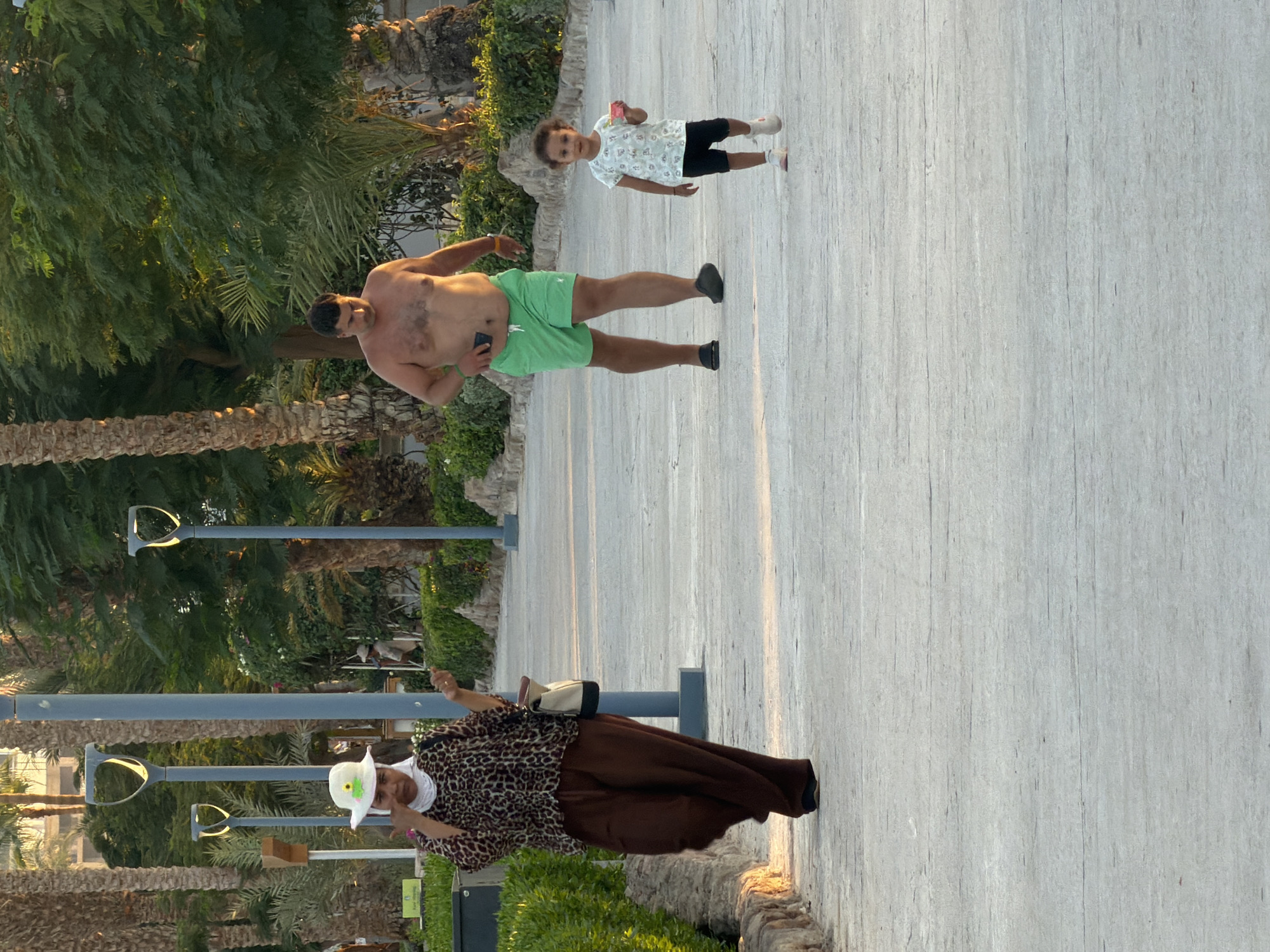The image shows a paved pathway in a park-like setting with palm trees and lush green foliage. The pathway is lined with stone borders and modern lampposts with decorative lights. In the foreground, a woman wearing a white hat, a leopard-print top, and brown wide-legged pants walks towards the left. She is carrying a handbag. Behind her and to the right, a man wearing green shorts and no shirt walks towards the right. Further to the right, a young child in a light-colored t-shirt and dark shorts walks towards the left, holding a small red object in their hand. The scene appears to be in a tourist area of Sharm El Sheikh, Egypt, given the tropical vegetation and the warm, sunny lighting which suggests an outdoor recreational space.