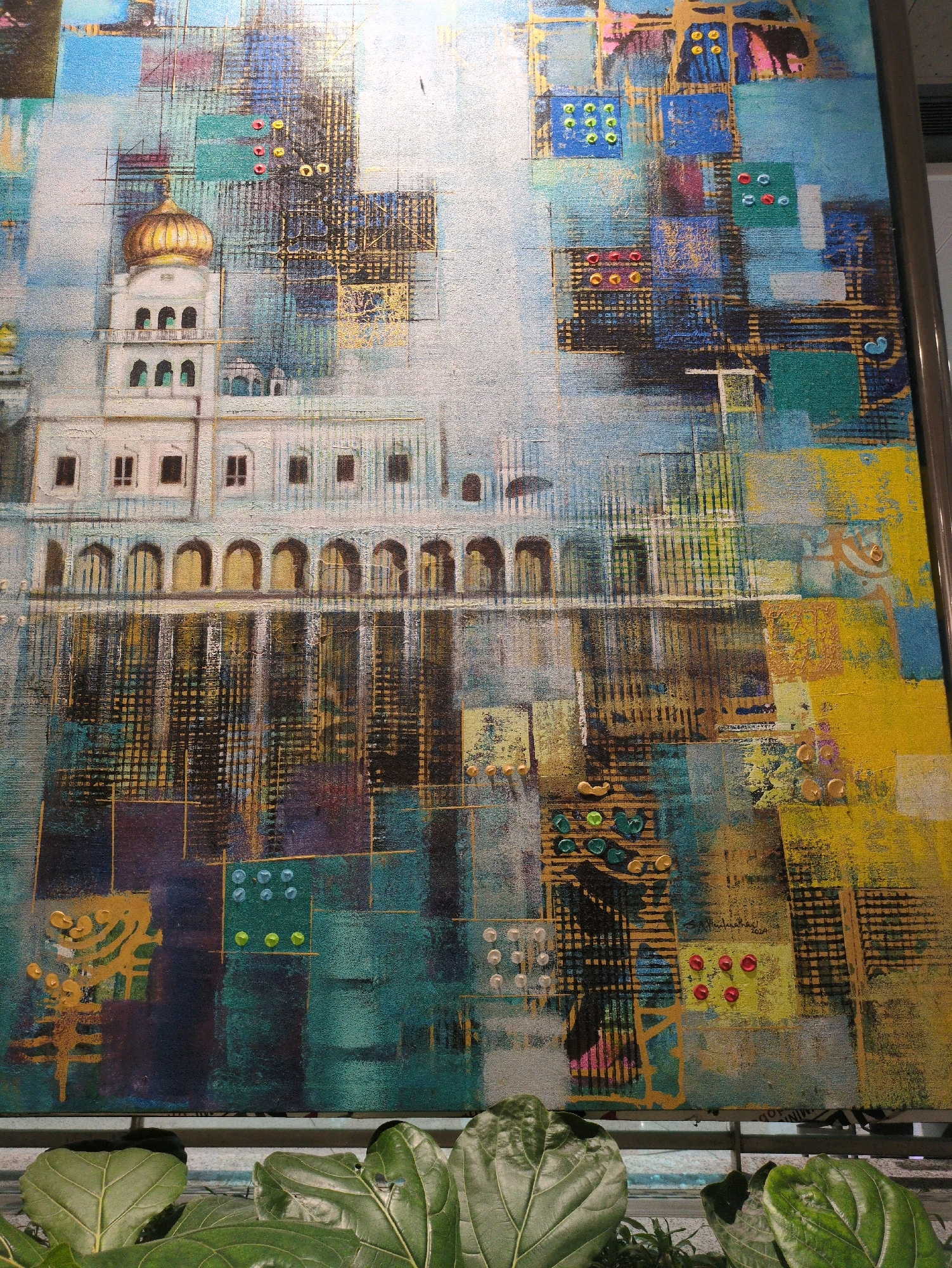 The image depicts an abstract painting with architectural elements, possibly inspired by a location in Delhi Cantonment, India. The artwork features a central white building with arched windows and a prominent golden dome, reminiscent of a historical monument or religious structure. The building is set against a backdrop of abstract geometric patterns, squares, and lines in various shades of blue, yellow, green, and black, creating a textured and layered effect. Small, circular embellishments in red, yellow, and green are scattered throughout the composition, adding a pop of color and dimension. The lower portion of the painting shows a reflection of the building in water, rendered with dark, vertical brushstrokes and streaks of gold. In the foreground, lush green leaves of a plant are visible, partially obscuring the bottom of the artwork and suggesting an indoor setting or a display near foliage. The overall impression is one of vibrant and dynamic art, blending representational and abstract styles.