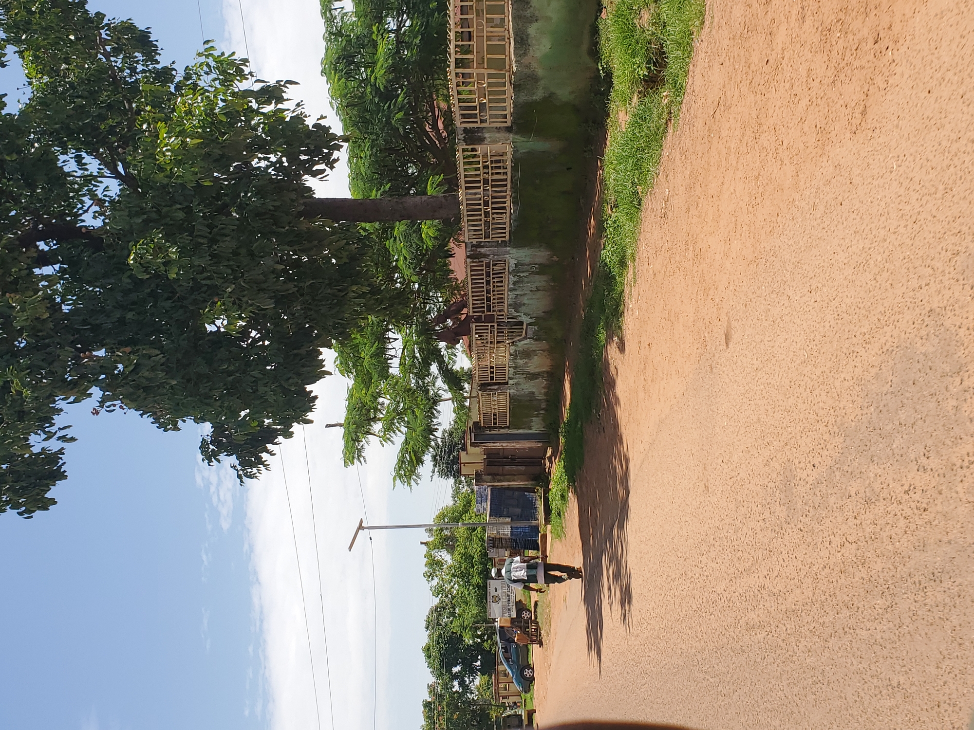 The image depicts a street scene in Pankshin, Nigeria, under a bright, partly cloudy sky, suggesting it is daytime. The camera is oriented vertically, making the scene appear sideways.
The foreground is dominated by a dusty, reddish-brown dirt road. To the left of the road, a line of buildings with weathered walls and fenced fronts runs along the side. Trees with dense green foliage are interspersed with the buildings, casting long shadows across the road. The trees appear to be of a tropical variety.
Further down the road, several objects are visible. A person is walking on the road, carrying what appears to be a large bundle on their head. There are a few vehicles, including what looks like a light blue car and a motorcycle. A lamppost stands on the side of the road, with electrical wires extending from it. Signs are also visible on some of the buildings, though the text is not legible in detail. One sign appears to be a white rectangular sign with some lettering. The overall impression is that of a typical street scene in a Nigerian town or city, with a mix of residential or commercial buildings, vegetation, and human activity. The weather appears to be warm and clear, conducive to outdoor activities.