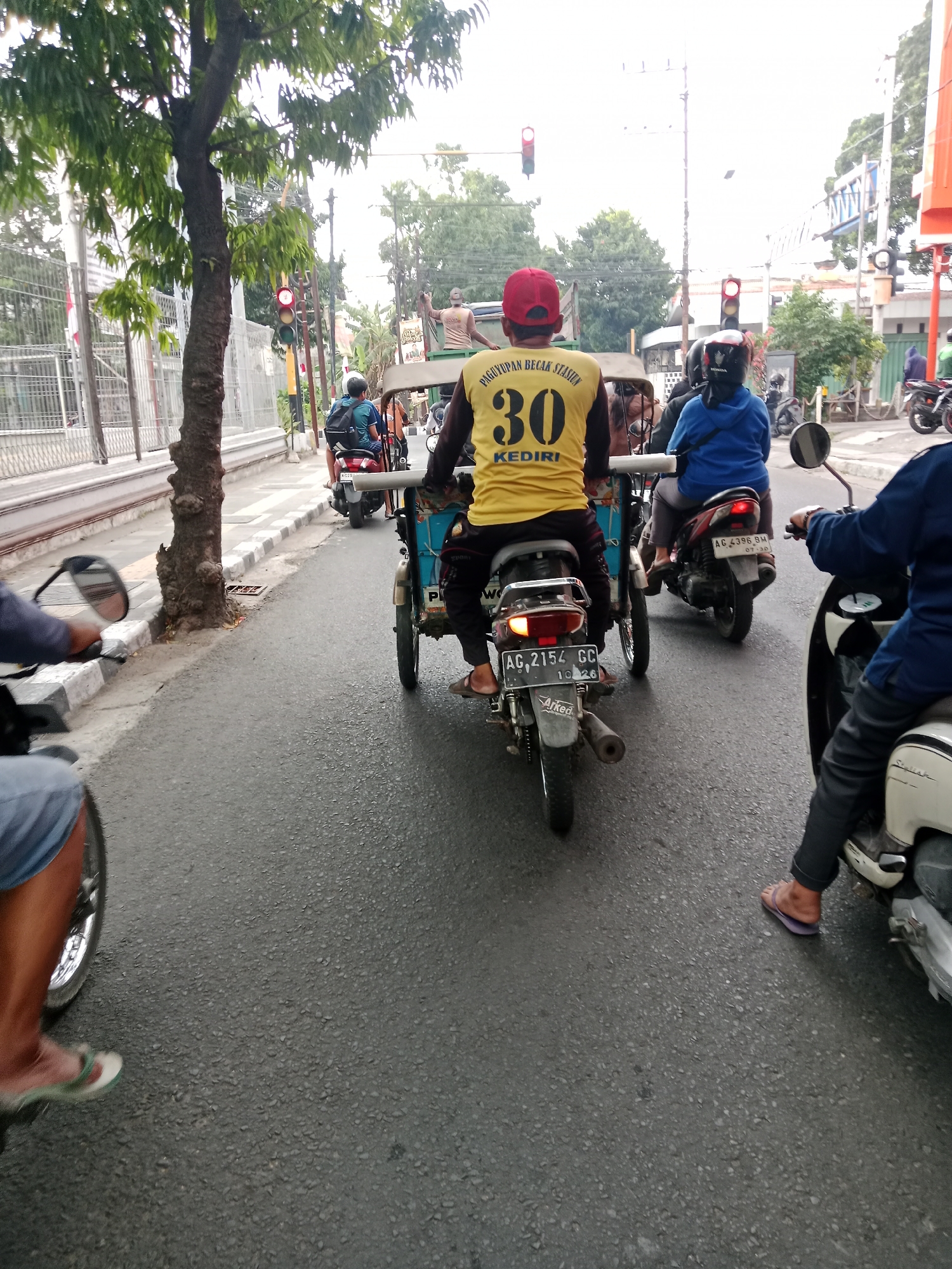 A medium shot captures a street scene in Kediri City, Indonesia, with several motorbikes and a pedicab moving in traffic. In the foreground, a person wearing blue jeans and sandals is on a motorbike, their legs and feet visible. Behind them, a pedicab with a driver wearing a yellow t-shirt and a red cap is prominent. The t-shirt has "PINGUYOAN BECAK KEDIRI" printed on the back, along with the number "30" and "KEDIRI". The pedicab has a license plate AG 2154 GC. To the right of the pedicab, another motorbike with a rider wearing a blue jacket is visible, with license plate AG 4395 BM. In the far right, a portion of another motorbike and a person wearing dark pants and sandals can be seen. The street is paved with asphalt, and there are trees and traffic lights in the background, indicating an urban environment. The sky is overcast.