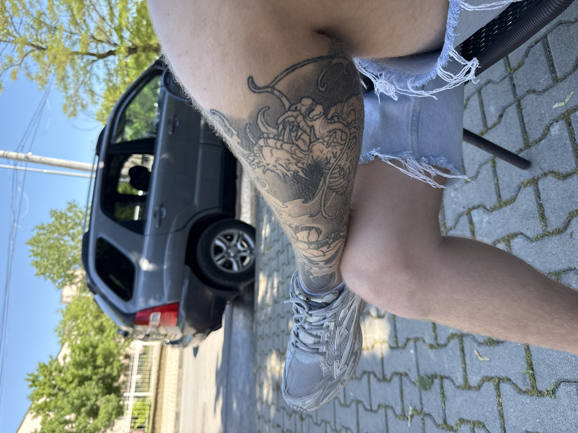 A medium shot shows a person sitting on a chair with their legs crossed. The person has a dragon tattoo on their right thigh, and they are wearing cutoff denim shorts and gray sneakers. In the background is a gray SUV parked on a paved surface. Trees and a blue sky are visible in the background. The photo was taken outdoors, possibly in Elhovo, Bulgaria.
