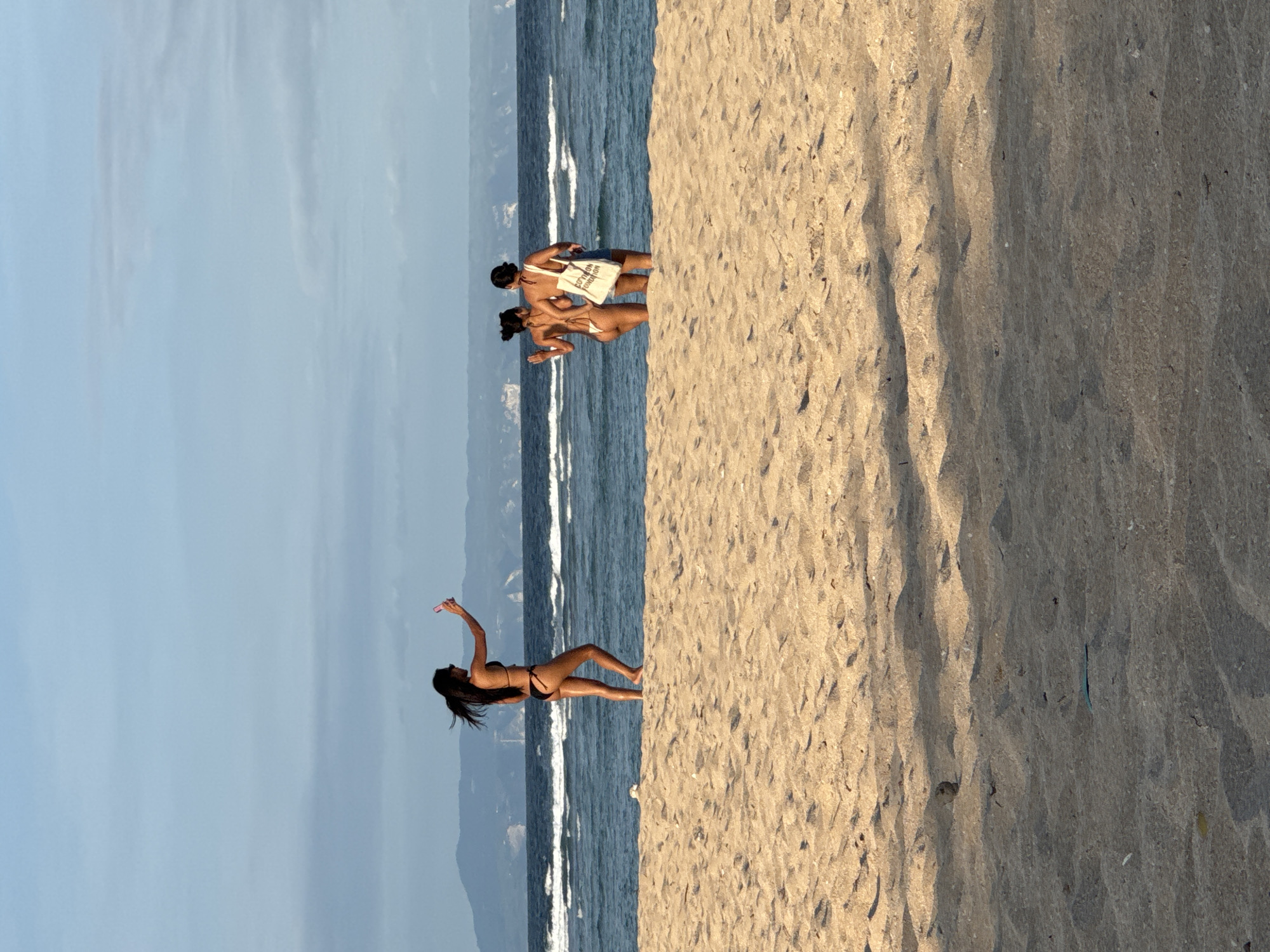 The image shows a beach scene, likely in Kuta Selatan, Indonesia, based on the visual characteristics of the beach and the presence of multiple individuals on the beach, some wearing swimwear. The beach is sandy and takes up the right half of the image. The water is on the left half of the image and there are several people in the foreground; one person is standing alone taking photos while another group is standing by the water. The background consists of the sky and land.