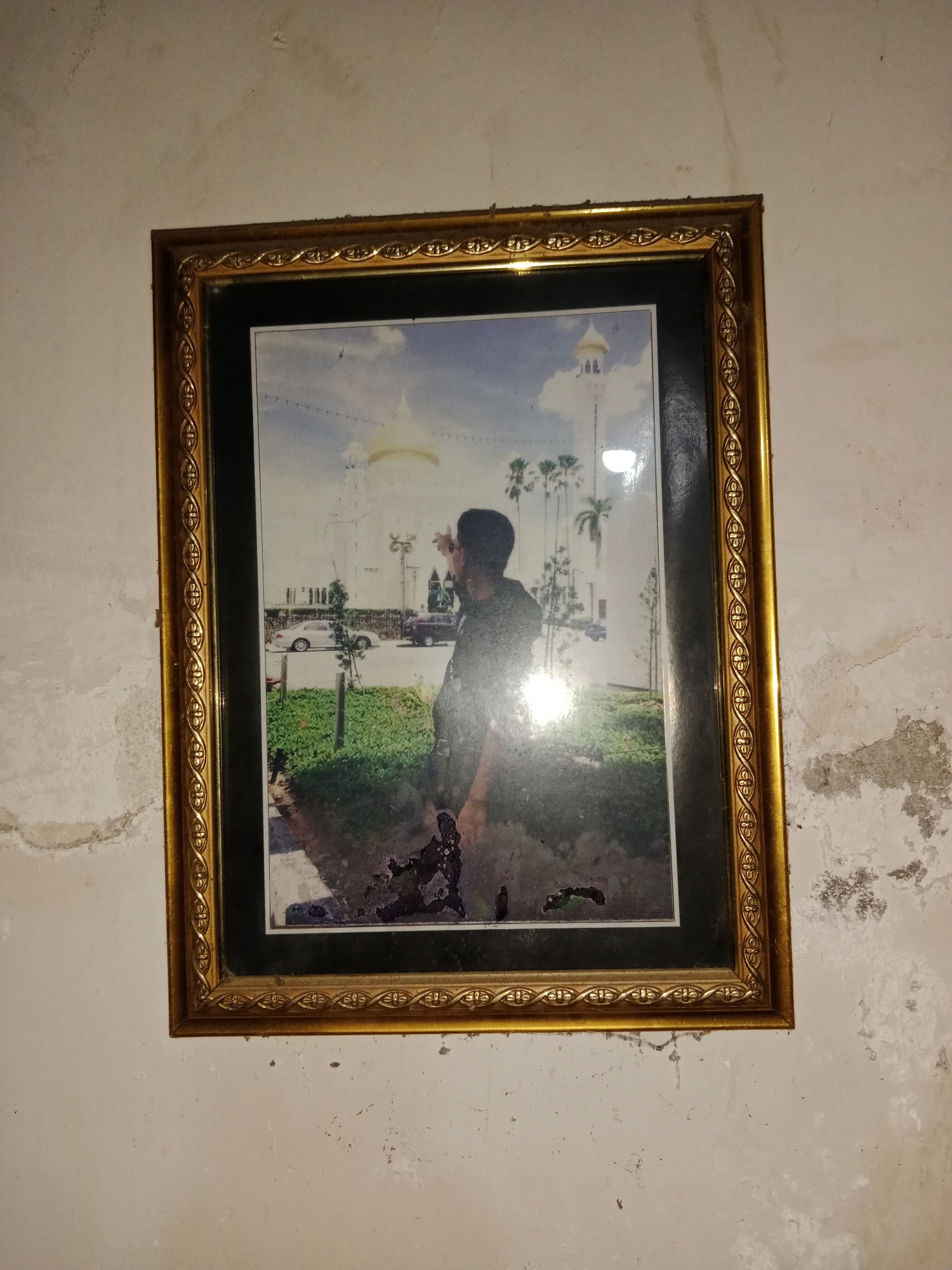 A framed photograph hangs on a weathered, textured wall. The photograph depicts a man standing outdoors, pointing towards a building with a golden dome and tall minarets, possibly a mosque, in the background. Several palm trees are also visible. A white car and a darker vehicle are parked in front of the building. The image itself shows signs of damage, with peeling and discoloration, particularly in the bottom right corner. The wall surrounding the frame is marked with stains and has a rough, uneven surface. The overall impression is of an aged and perhaps neglected space. The presence of a mosque and palm trees suggests a location in a tropical or subtropical climate. Ringinrejo, Indonesia, is a town located in East Java, a region known for its mosques and cultural heritage, consistent with the visual cues in the photograph.