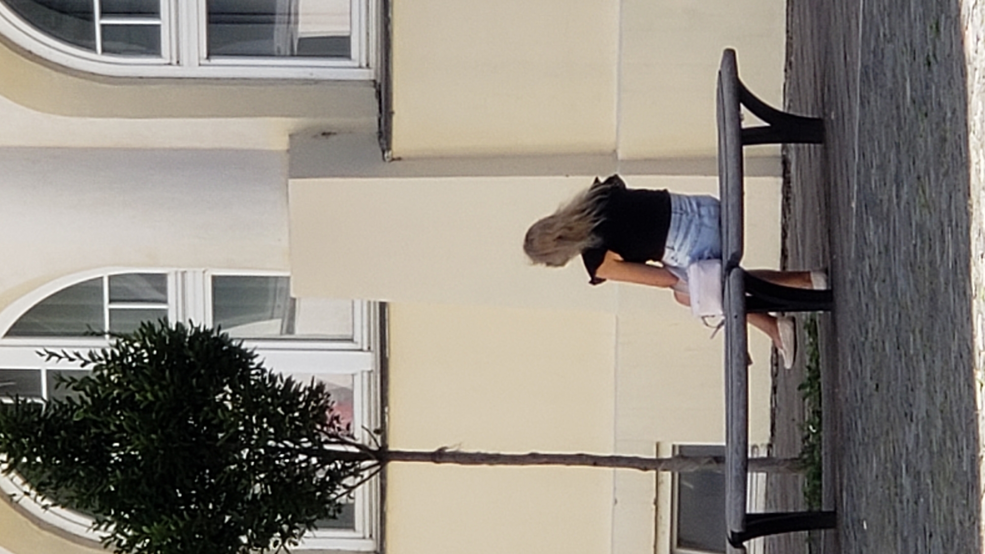 A blonde woman is seated on a park bench facing away from the camera. She is wearing a black ruffled top and light blue denim shorts. A light pink purse rests beside her on the bench. The bench is dark gray and appears to be made of metal and wood. Behind the bench is a building with arched windows. To the left of the bench is a thin, leafy tree. The ground in front of the bench is paved with dark cobblestones. The scene appears to be in the city of Prešov, Slovakia.