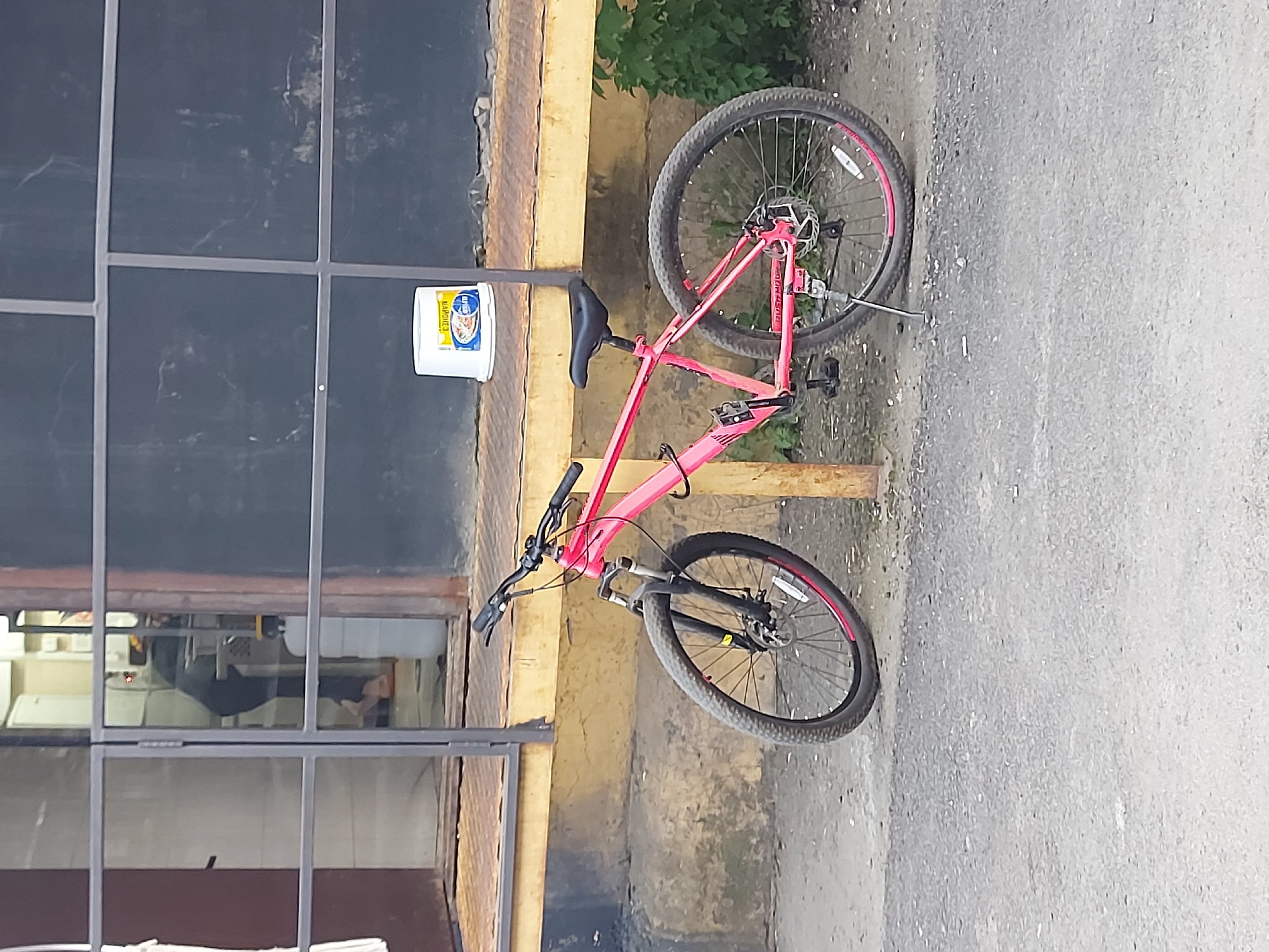 The image shows a pink mountain bike leaning against a yellow guardrail in an urban setting. A white plastic bucket with blue writing on it is placed on the guardrail next to the bike's seat. A building with a glass-paned window is visible in the background, reflecting interior scenes. The setting appears to be outdoors, with a paved surface. Based on the image, there are no indications of this location being Izhevsk, Russia. More data is required to make that determination.