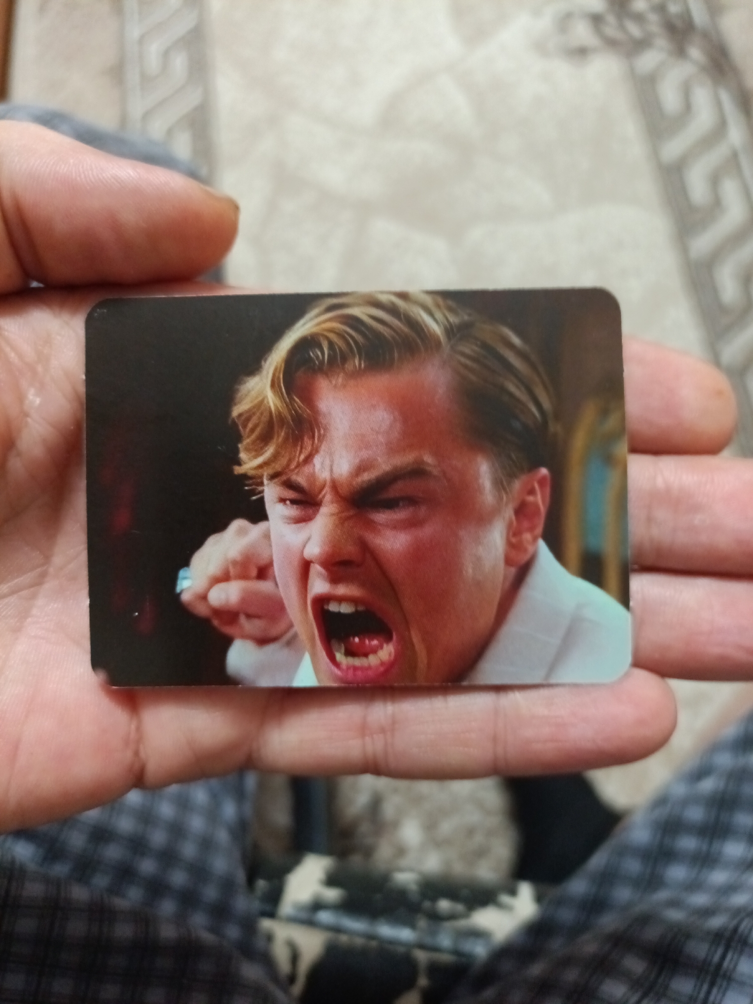 A close-up shot shows a person's hands holding a rectangular card featuring a screenshot of Leonardo DiCaprio from "The Great Gatsby." DiCaprio's face is contorted in an angry expression, mouth open in a shout, with his right fist clenched and raised towards the left side of the frame. The background of the card is dark, with a hint of a golden, ornate interior. The hands holding the card appear to be positioned over a patterned surface, possibly a blanket or upholstery, with a faint floral design visible. The lighting suggests an indoor setting. The image does not provide any information or context related to the city of Nyagan, Russia.
