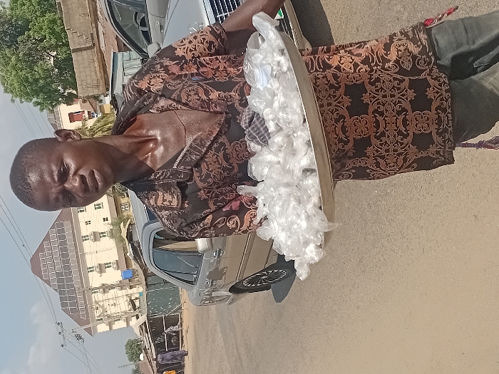 A dark-skinned man stands on a sunny street, holding a silver tray filled with small, likely bagged items. He wears a patterned shirt with rolled-up sleeves, dark pants, and appears to be perspiring. A silver car is positioned beside him. In the background, buildings are visible under a clear sky. The scene suggests street vending or sales activity in an urban setting.