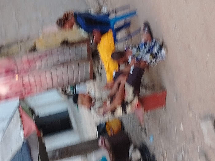 The image depicts three individuals in an outdoor setting within the city of Orji, Nigeria. An adult male, wearing a dark cap and a light-colored patterned shirt, is seated on a red stool or low bench in the foreground-left, with his legs extended. To his right, an adult female wearing a dark blue top is seated on a blue plastic chair; a yellow fabric is draped over her lap area. Between the two adults, a child in a patterned blue and white outfit is seated directly on the dirt or sandy ground. The individuals appear to be resting or engaged in a casual outdoor gathering. In the background, various structures are visible, including a multi-colored tarp or awning on the upper left and sections of corrugated metal and light-colored walls. A container is present on the ground to the left of the male, and a white bottle is partially visible behind him. The entire scene is characterized by significant motion blur.