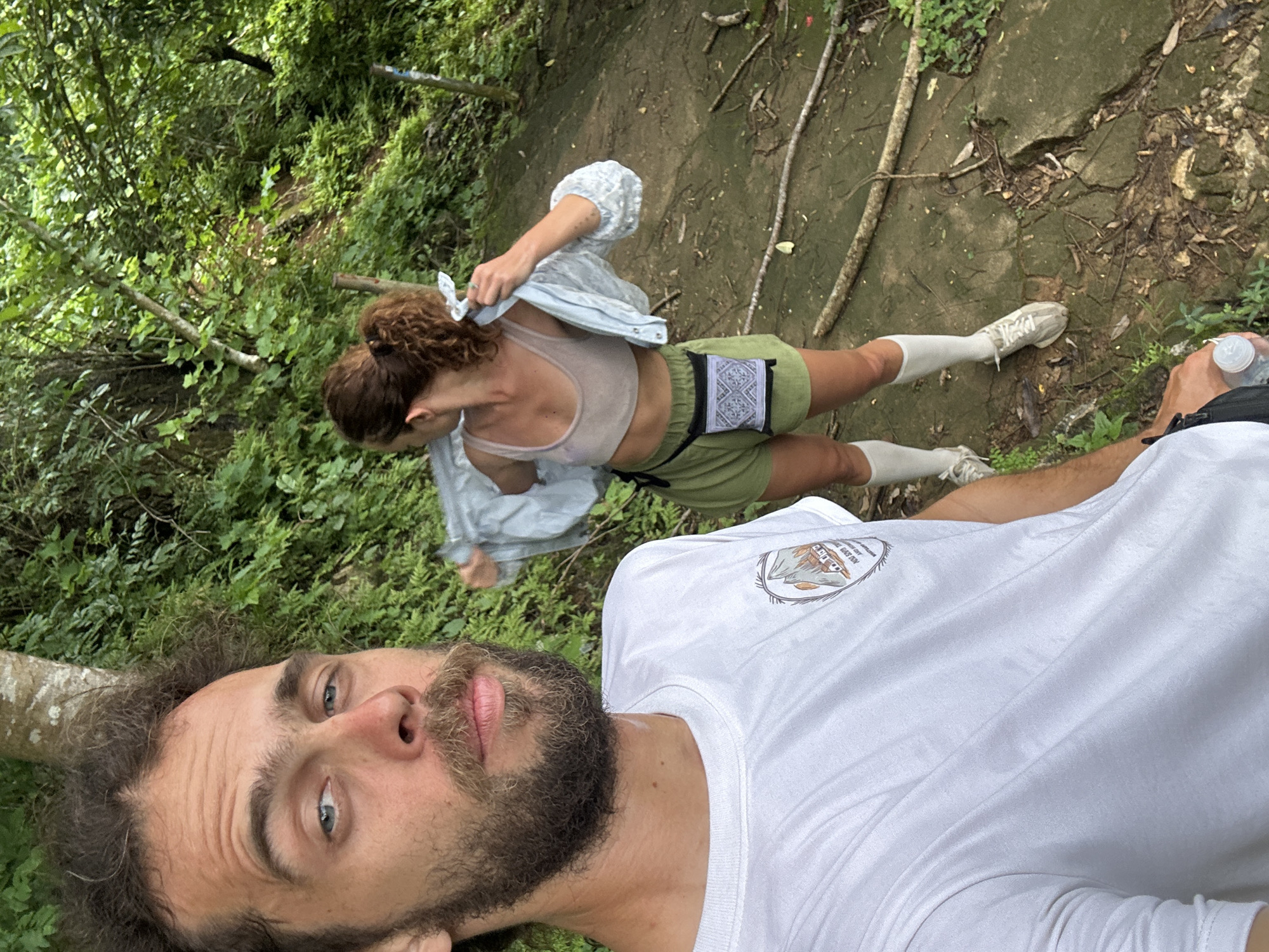 A man in a white t-shirt and a woman in a light-colored athletic top and green shorts are posing for a selfie on a hiking trail in Mai Chau, Vietnam. The man is in the foreground, facing the camera with a beard and curly hair. The woman is in the background, turning away from the camera and adjusting her clothing. The trail is made of concrete and dirt, with lush green foliage surrounding it. The weather appears to be overcast, with a humid atmosphere suggested by the man's damp skin.