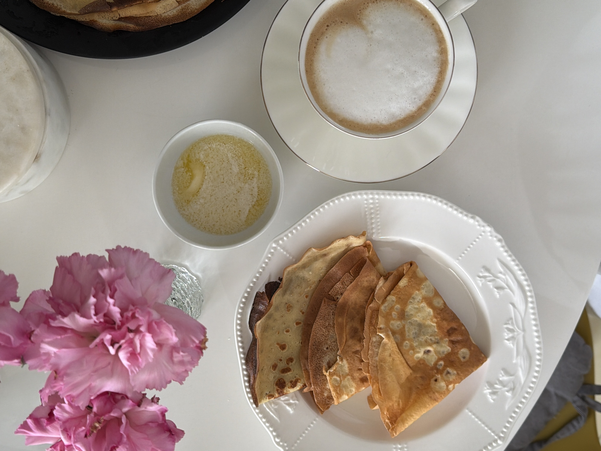 This image depicts a breakfast scene on a white surface. A plate of folded crepes sits next to a small bowl containing what appears to be melted butter and granulated sugar. Adjacent to these items is a cup of coffee or cappuccino on a saucer. In the lower left corner is a vase holding pink flowers. The image may be a photograph taken in a private residence or cafe in Khimki, Russia but this is not possible to determine with the information given.