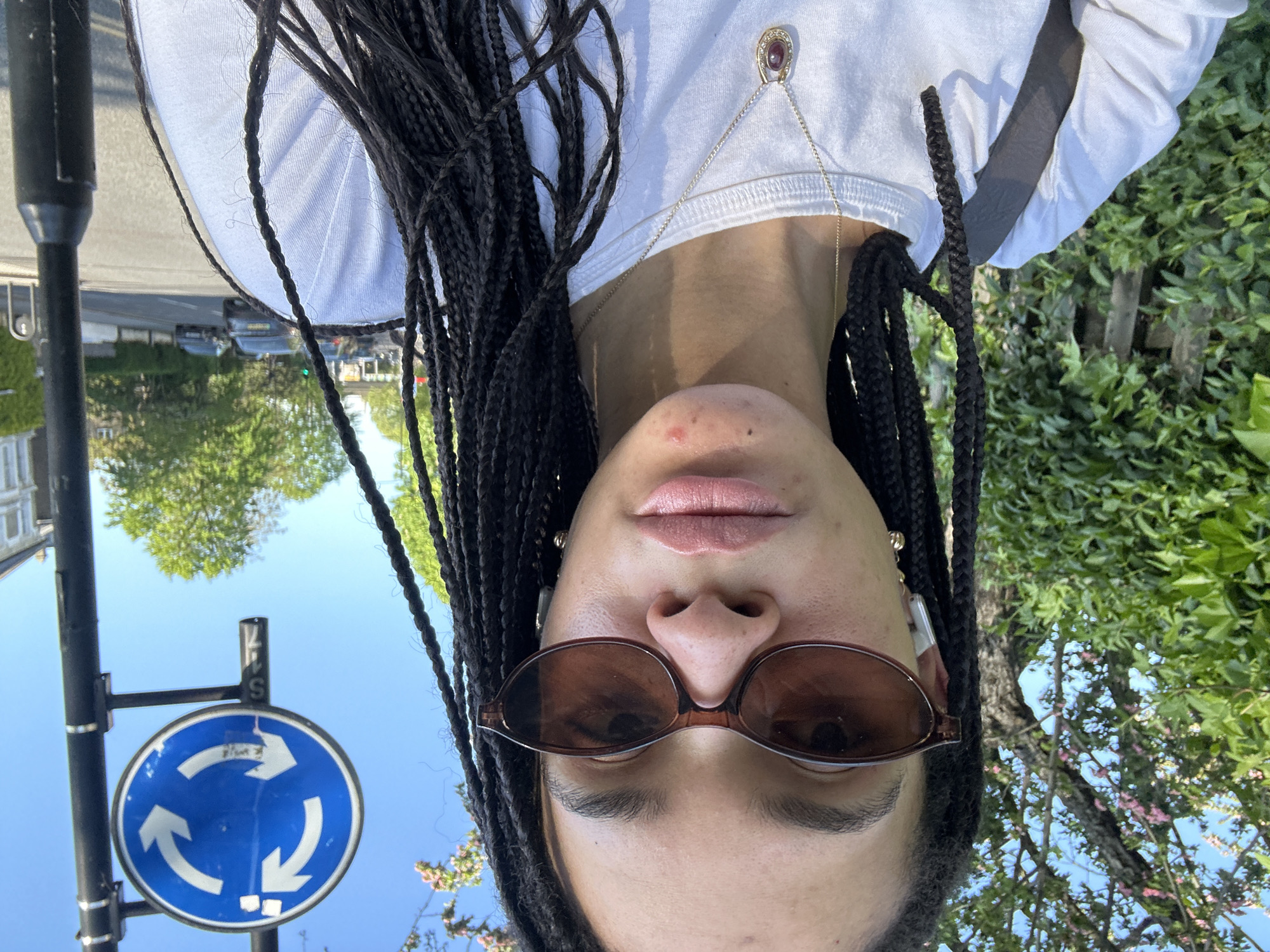 A selfie of a woman with long braids wearing sunglasses and a white shirt. She is standing on a street in London, United Kingdom. A blue roundabout sign is visible in the background. Green trees and buildings line the street. The sky is blue.