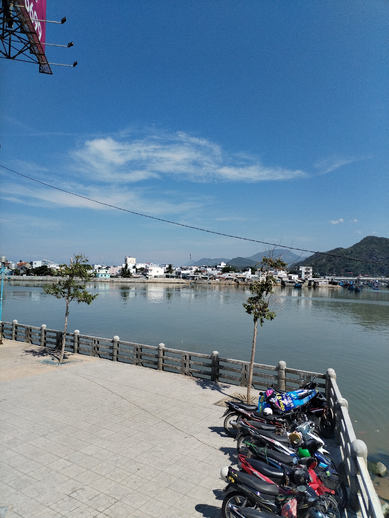 The image shows a waterfront scene in Bac Nha Trang Ward, Vietnam. A paved promenade lined with a concrete balustrade borders a body of water. Several motorbikes are parked along the balustrade. Two small trees grow on the promenade. Across the water, a town is visible, backed by hills. The sky is blue with scattered clouds. A billboard structure is partially visible in the top left corner of the image.