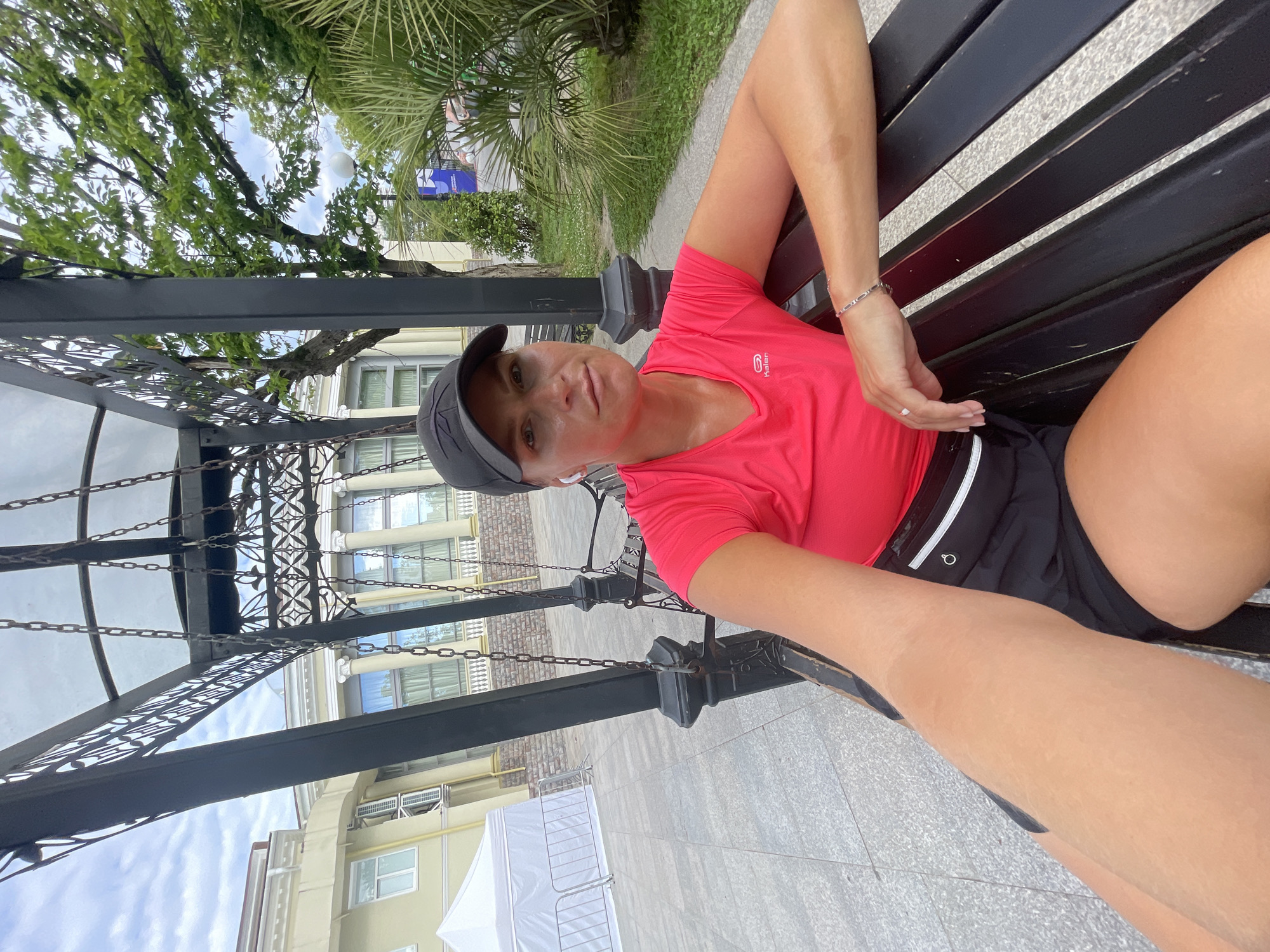 A woman sits on a black park bench in front of a building with classical architecture, which is located in the city of Sochi, Russia. The woman is wearing a bright pink t-shirt, black shorts, a black baseball cap, and white earbuds. She has fair skin and is wearing light makeup. Her right arm is resting on her leg, and she is looking towards the camera. The bench is made of dark wood and is part of a swing set structure. In the background, there is a building with columns and a white tent structure with a fence in front of it. There are also palm trees and other greenery visible. The sky is cloudy. The image appears to be a selfie.