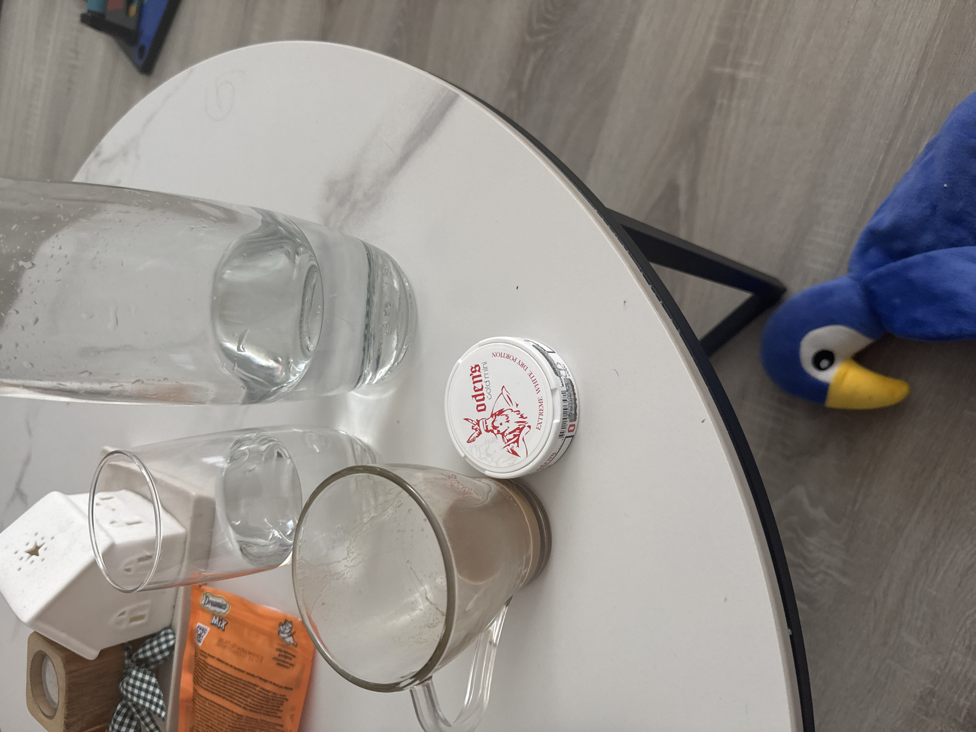 The image depicts a white marble-patterned round table indoors. On the table are a clear glass water bottle lying on its side, a clear glass lying on its side, an empty glass mug with a handle, a white and tan ceramic object with a star cutout, a bag of Dreamies cat treats, a black and white checkered ribbon, and a white circular tin of Oden's snus. A blue and yellow plush toy is partially visible on the right edge of the table. The setting is indoors, likely in a private residence. There is no evidence to suggest the location is in Füssen, Germany.