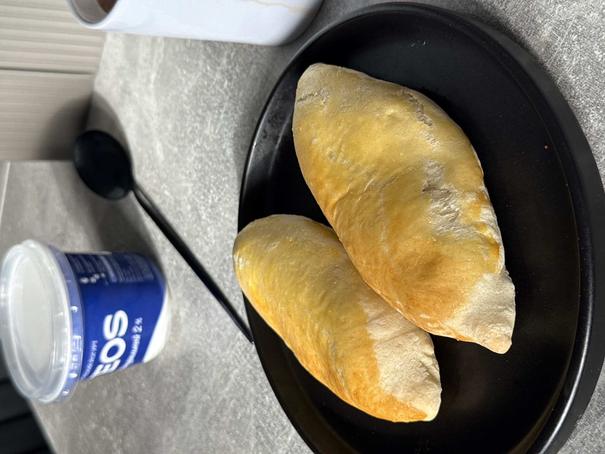 A close-up shot shows two golden-brown, oval-shaped baked goods, possibly pies or pastries, resting on a black plate. The plate is situated on a textured gray surface, resembling a countertop. In the background, a partially visible white mug with some liquid and a dark spoon are present. To the left, a blue and white plastic container with a clear lid is also visible. The overall lighting suggests an indoor setting. There is no specific indication of the location being Lyubertsy, Russia.