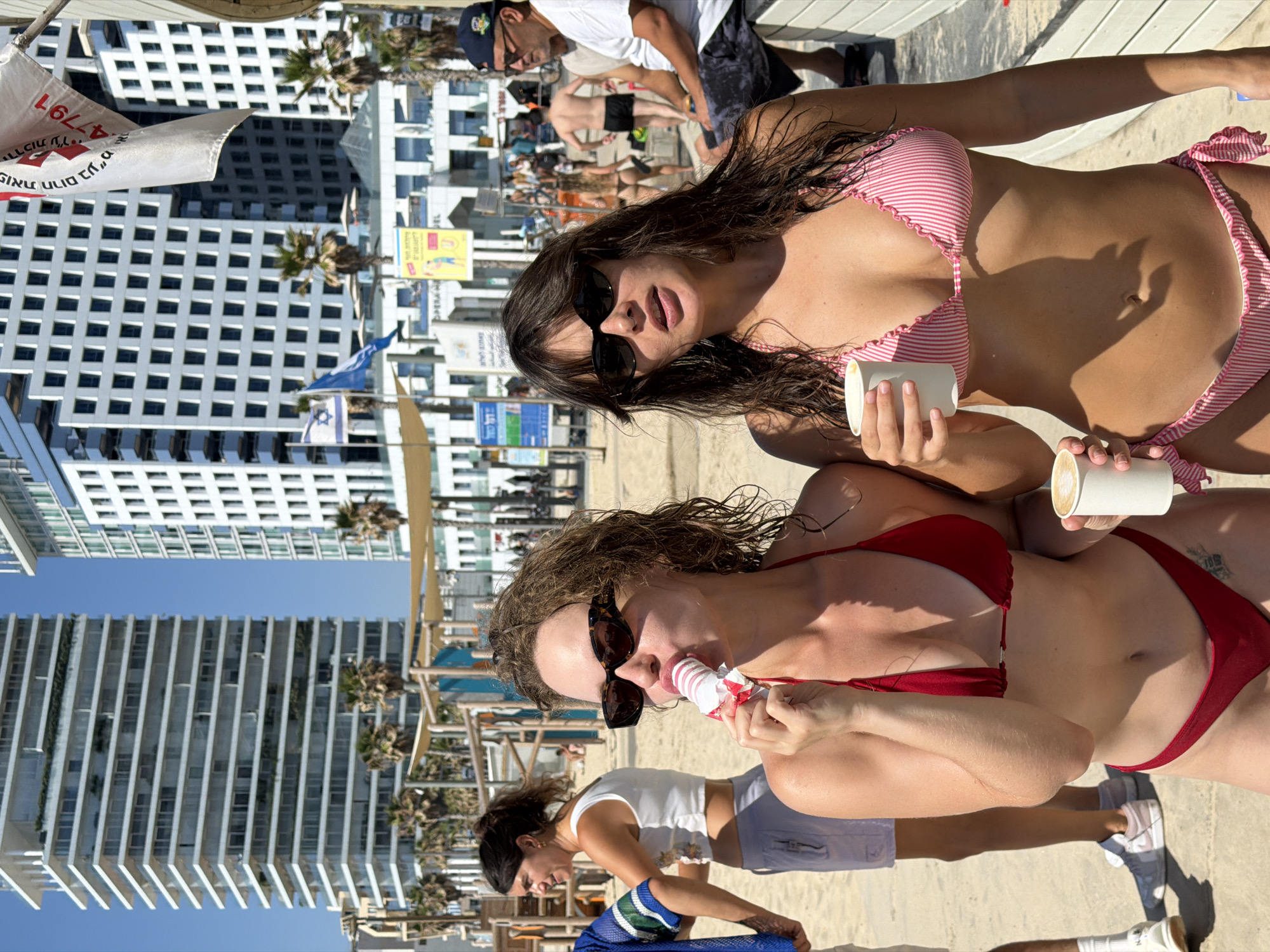 This is a medium shot photograph taken on a sunny day at a beach in Tel Aviv, Israel. In the foreground, two young women are featured. The woman on the left, with curly brown hair, is wearing sunglasses and a red bikini. She is holding a white popsicle and appears to be eating it. The woman on the right, also with brown hair, is wearing sunglasses and a pink and white striped bikini. She is holding a white cup. Behind them, a woman with dark hair pulled back is wearing a white crop top and grey shorts. In the background, there are tall modern buildings, palm trees, and beach amenities like umbrellas and chairs. Several other people are visible in the distance, some walking or sitting on the sand. A flag with Hebrew writing is also visible, fluttering in the breeze. The scene suggests a casual beach atmosphere, likely in the city of Tel Aviv due to the architectural style of the buildings and the beach setting.