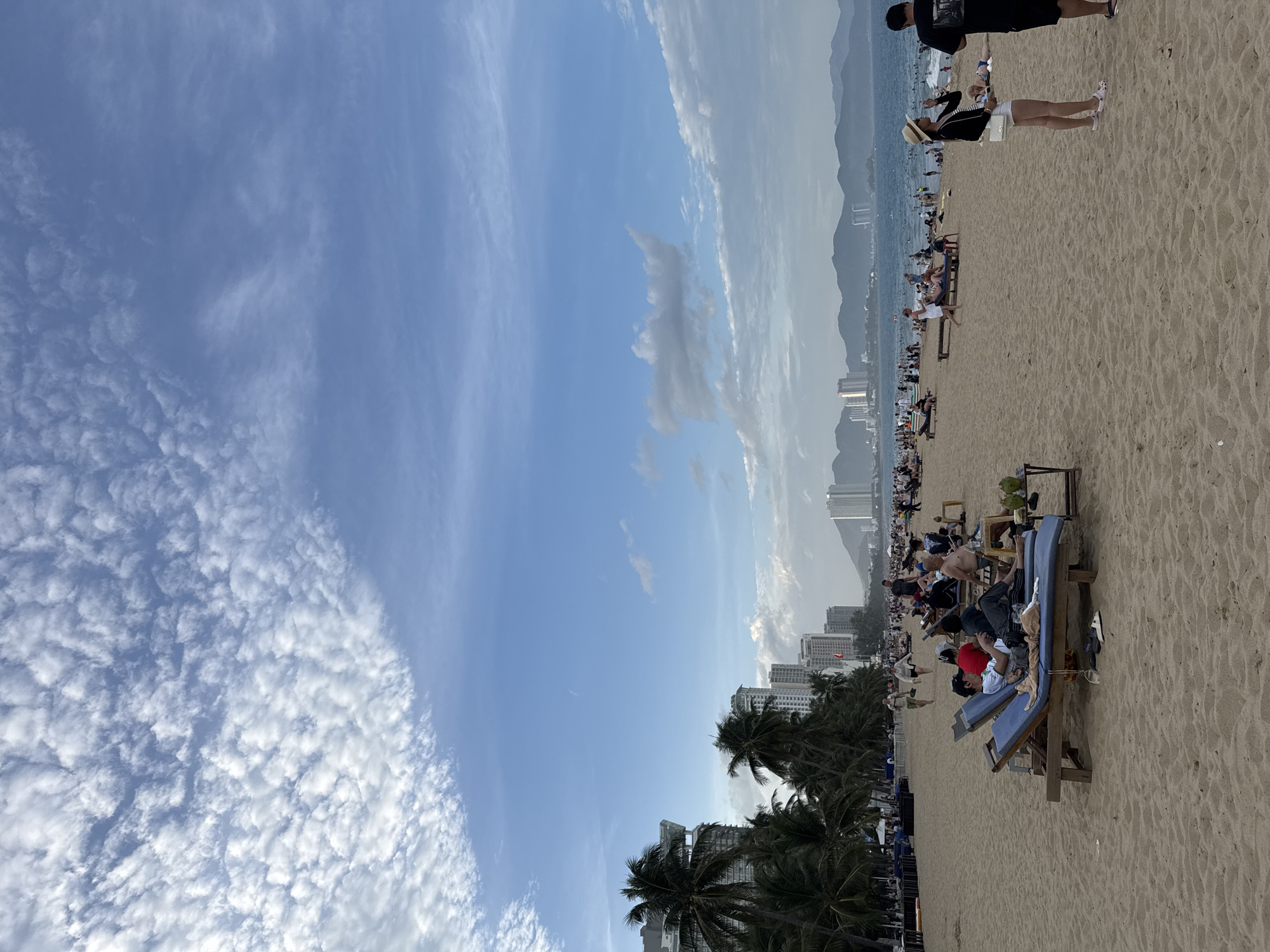 The image depicts a bustling beach scene in Nha Trang, Vietnam, likely Nha Trang Beach. The foreground is dominated by golden sand with numerous beachgoers relaxing on lounge chairs. In the mid-ground, the ocean stretches to the horizon, dotted with people swimming and engaging in water activities. A prominent line of tall buildings and hotels runs along the coastline, characteristic of Nha Trang's urban development. Palm trees are scattered throughout the scene, adding to the tropical atmosphere. The sky is a clear blue with scattered white clouds, suggesting fair weather. People are seen in various states of relaxation, some lying on loungers, others walking, and some gathered in groups. The overall impression is one of a vibrant tourist destination.