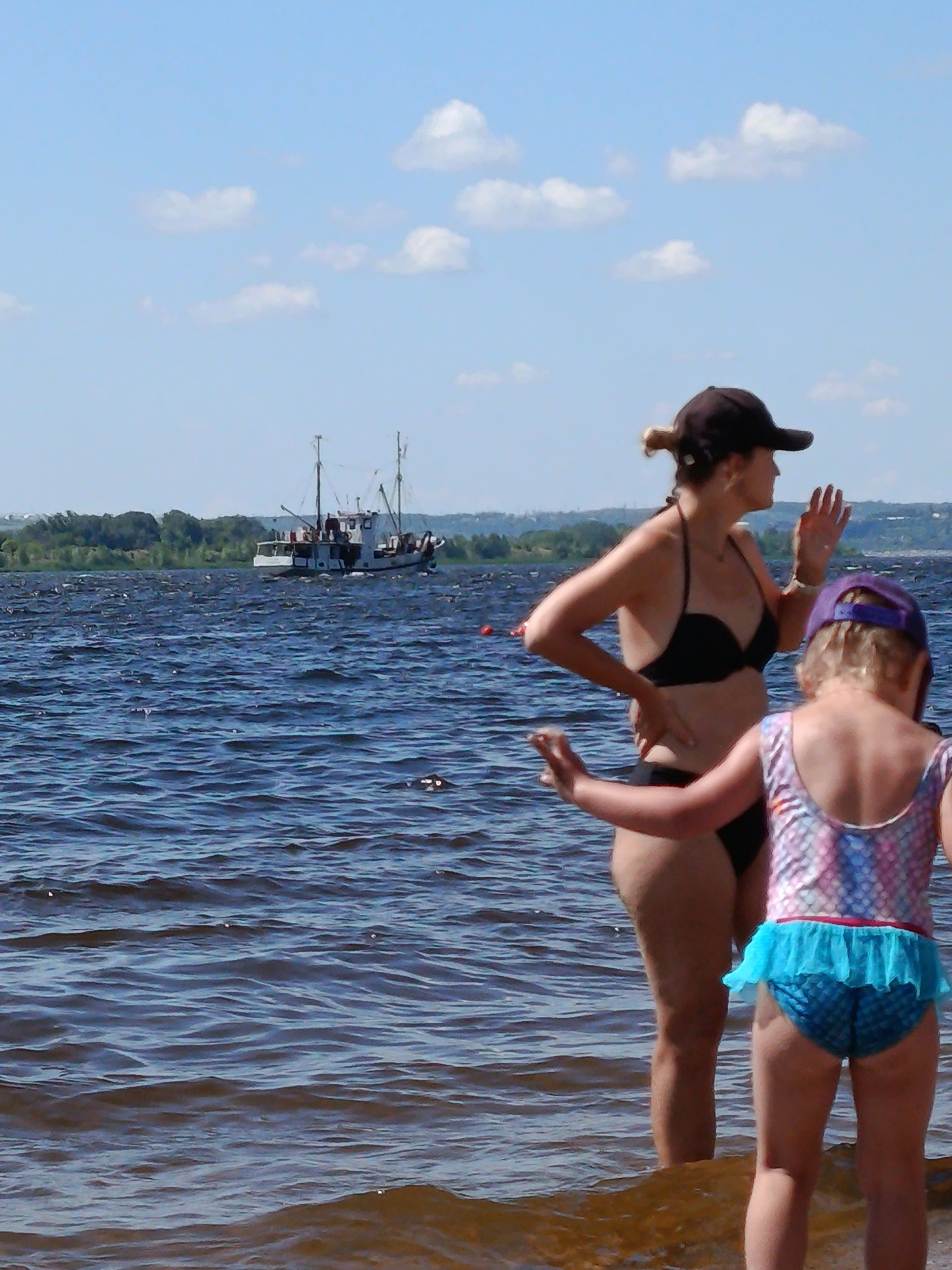 The image depicts two females, an adult and a child, standing in shallow water. The adult, wearing a black bikini and a baseball cap, is facing away from the camera, with her right hand raised in a waving gesture. The child, in a swimsuit with a mermaid-scale pattern and blue ruffled bottoms, is also facing away, with her arms extended forward.
In the background, a boat is visible on a wide body of water, with a tree-lined shore and a hazy skyline beyond. The sky is a clear blue with scattered white clouds, suggesting a sunny day. The water is choppy, with small waves indicating a gentle breeze. The scene appears to be a recreational area, likely a beach or a lakefront, suitable for swimming or boating. The overall mood is one of leisure and enjoyment on a summer day. No visible text is present in the image. The location context is Saratov, Russia.