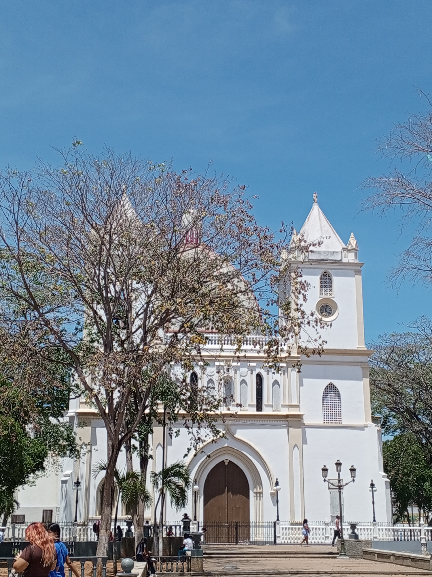 The image shows the Our Lady of the Valley Basilica, a large white church in Porlamar, Venezuela. The church has a tall, white tower with a clock on it, and a large brown wooden door. There are trees in front of the church, some of which are bare. People can be seen walking and sitting around the church, on the public square. The sky is blue.