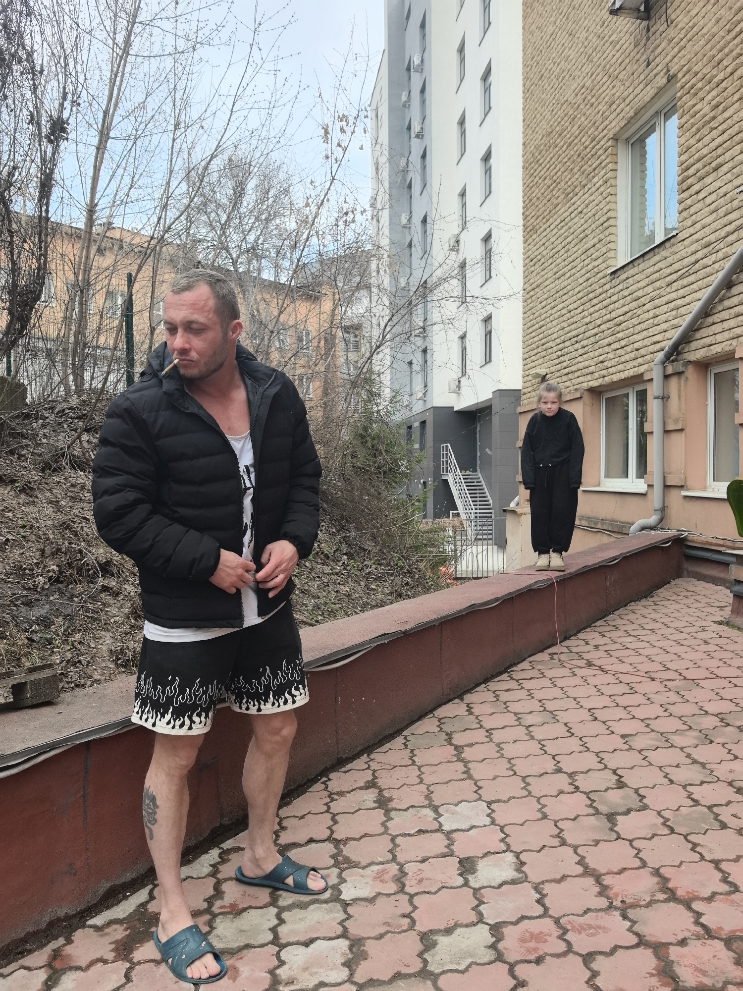 A man stands smoking on a brick-paved area with a low wall on his left and a building on his right. The man is wearing a black jacket, a white tank top, shorts with a flame pattern, and blue sandals. A girl wearing a black jumpsuit and standing on the low wall. Buildings of varying heights and colors are visible in the background, along with bare trees. The location is Nizhny Novgorod, Russia.