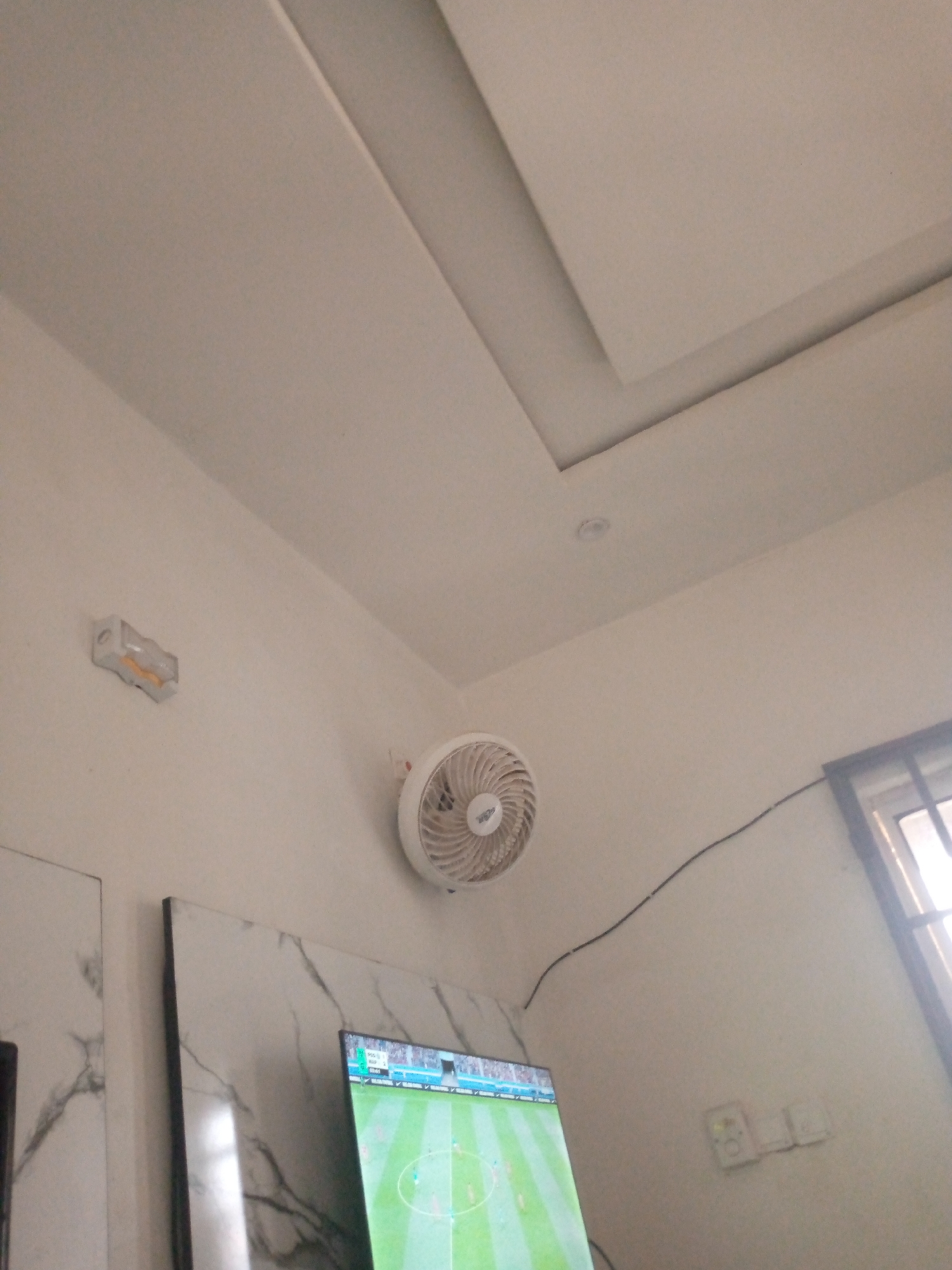 The image captures an interior view of a room, likely a living area, in Jalingo, Nigeria. The dominant feature is a white ceiling with recessed lighting and geometric detailing. Mounted on the white wall is a small, white electric fan, circular in design, with visible blades and a central logo. To the left of the fan, a wall-mounted light fixture, possibly an emergency exit sign, is visible. Below and to the right of the fan, a television screen displays a soccer match, with players on a green field. To the left of the television, a portion of a marble-patterned panel is visible. In the far right of the image, a window with a black frame allows light to enter, and below it are three electrical outlets. A black cable snakes across the wall from the fan towards the window. The overall scene suggests a domestic environment.